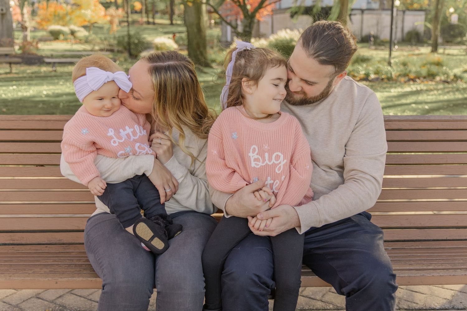 A family wearing coordinating pink and gray outfits sits together on a wooden bench in an outdoor setting.