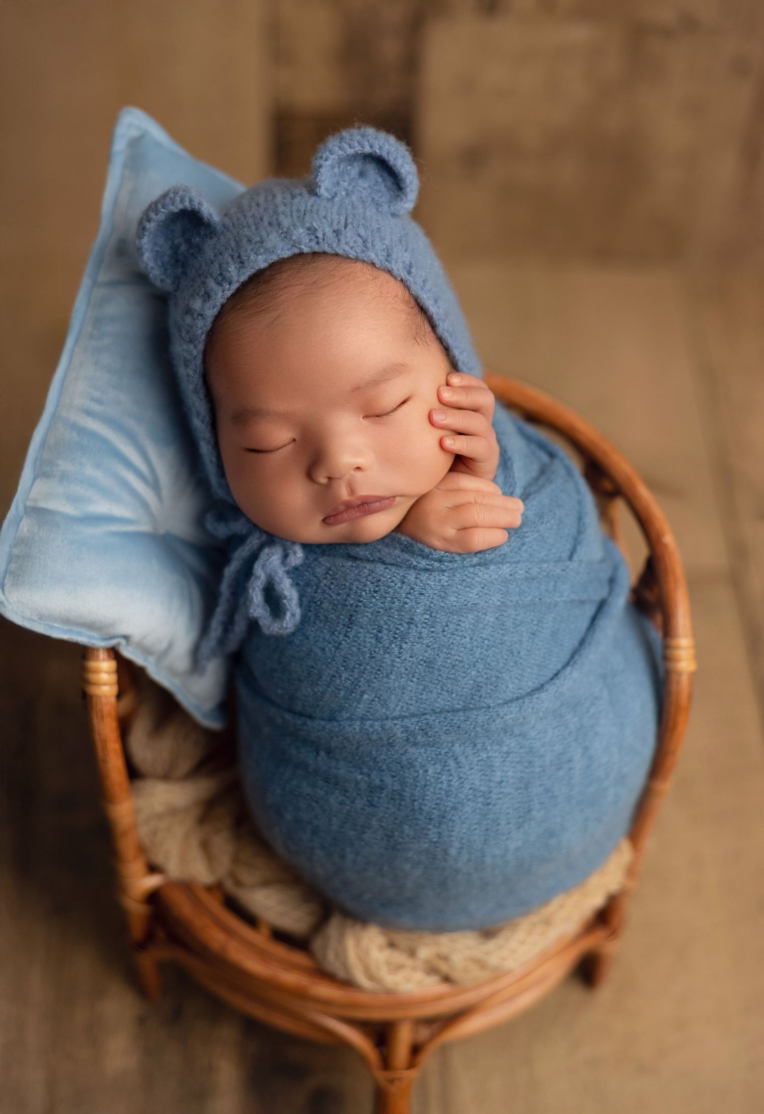 Sleeping newborn wrapped in soft blue swaddle wearing bear ear hat rests peacefully in wicker basket with blue pillow.