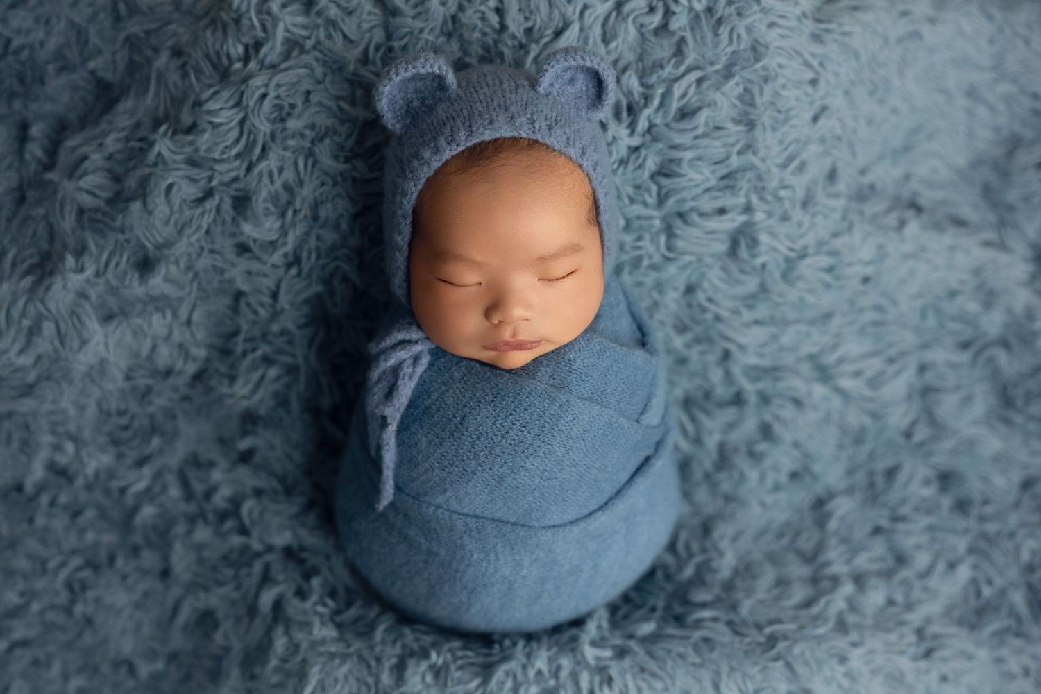 A sleeping baby lies on soft blue textured fabric in a professional newborn photography session.