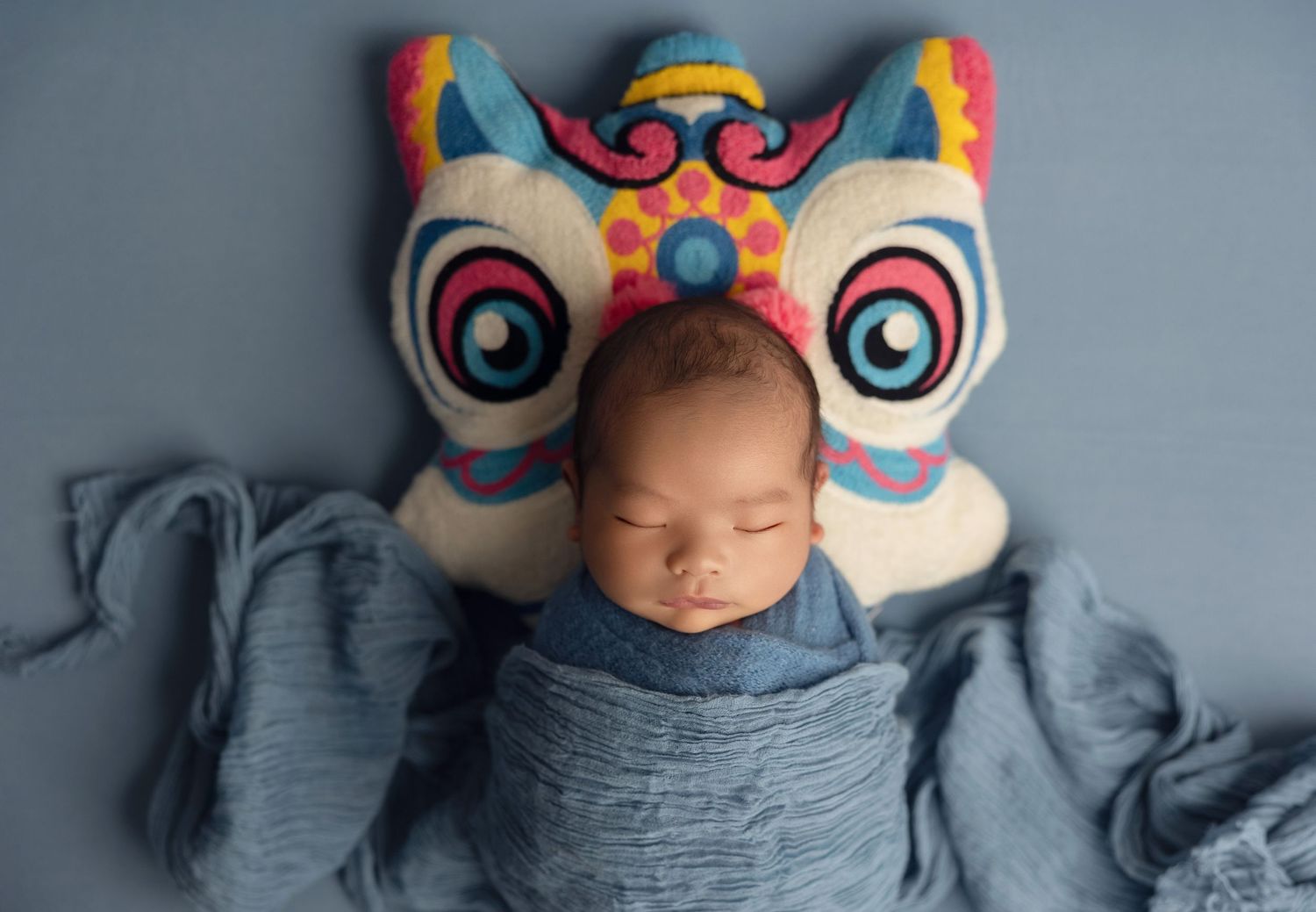 A sleeping baby wrapped in gray swaddle blanket with colorful owl pillow decoration above their head.