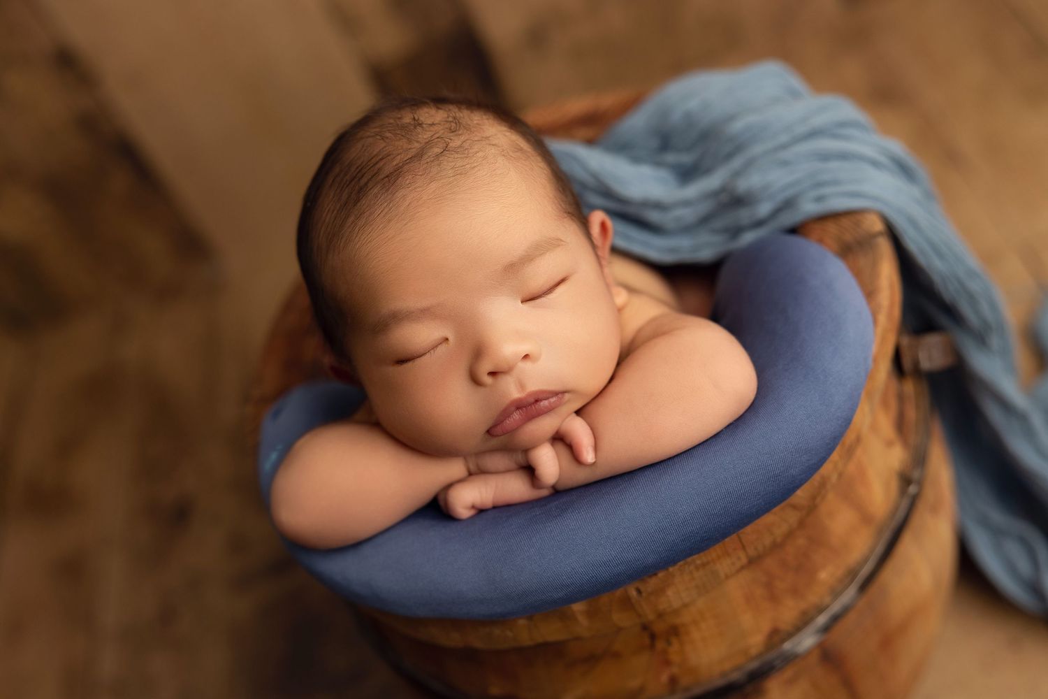 Sleeping newborn baby rests peacefully on a blue pillow with brown blanket in soft natural lighting.