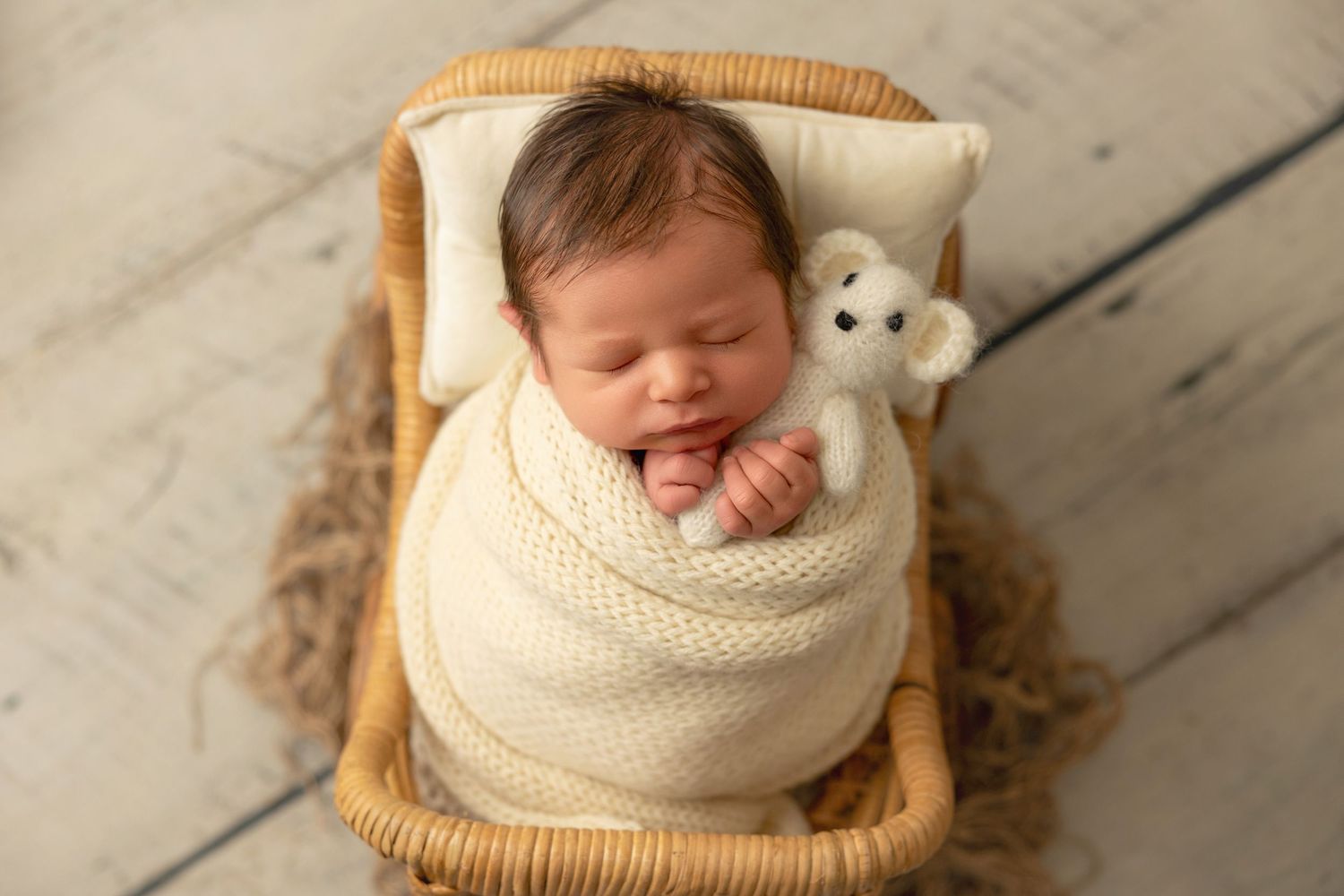 Sleeping newborn wrapped in cream knit blanket rests in wicker basket with small stuffed bear toy.