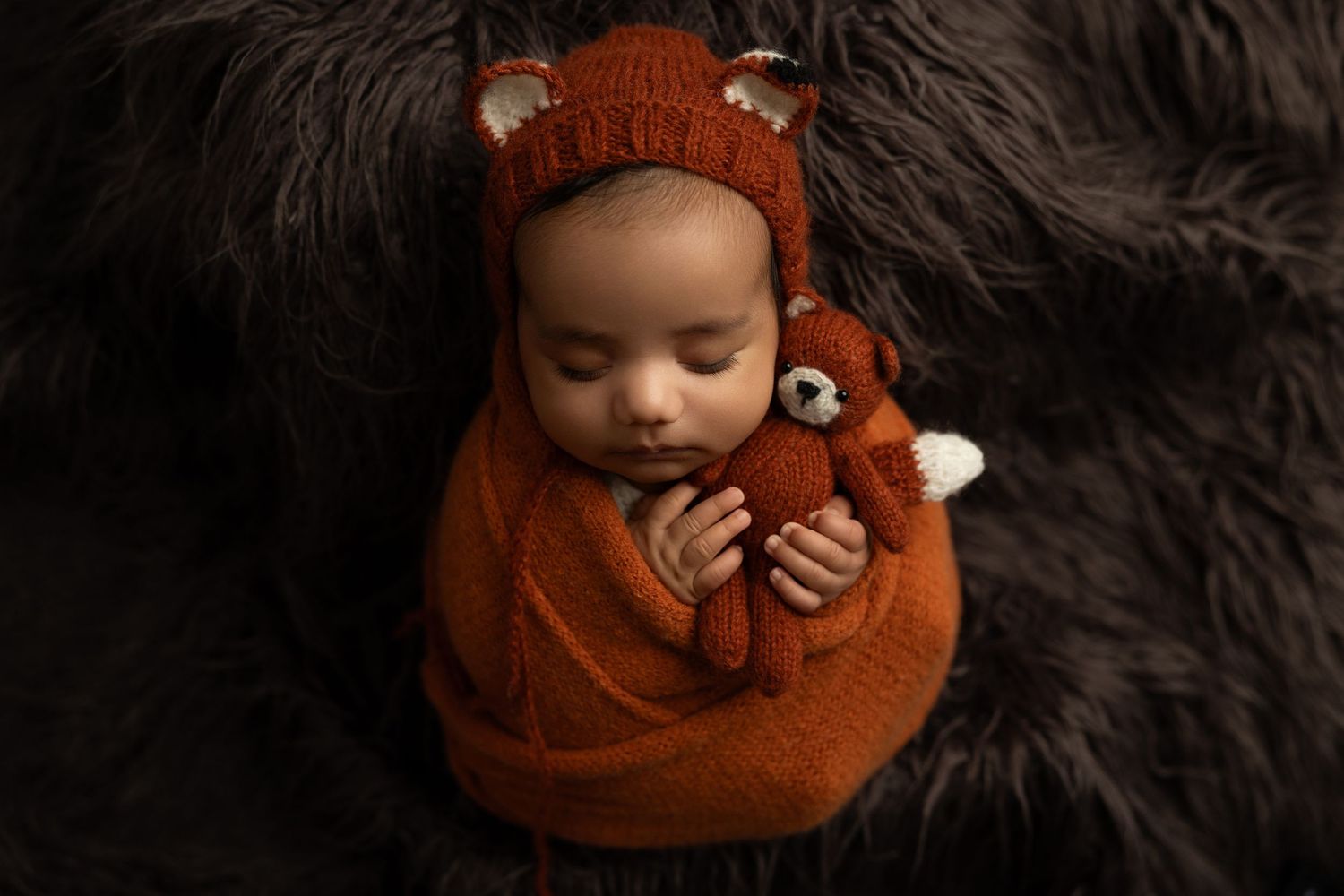 A baby wearing a cozy brown knit outfit lies on a dark furry blanket while sleeping peacefully.