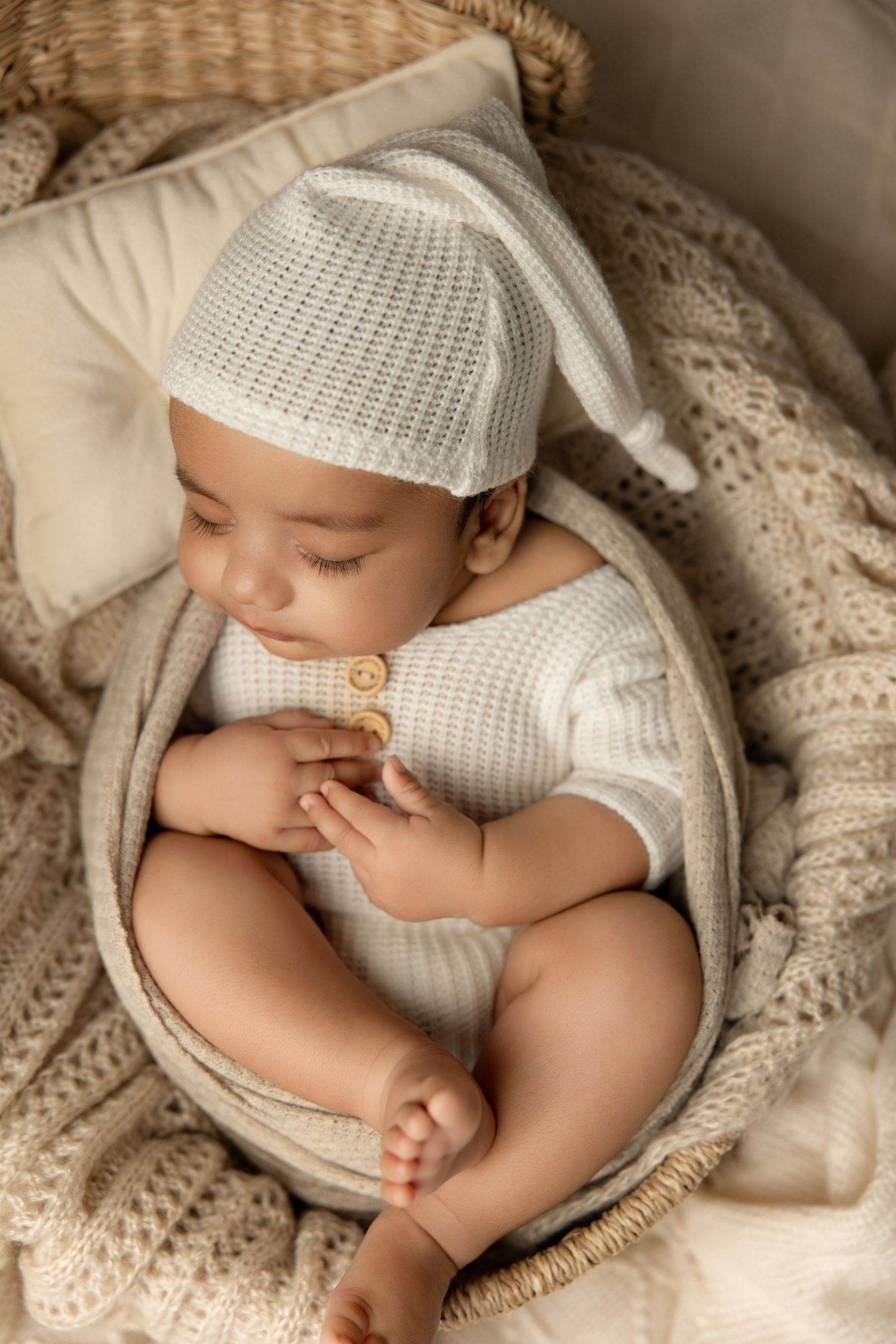 A sleeping baby wearing a white knit outfit rests peacefully on cream-colored blankets in a cozy basket.