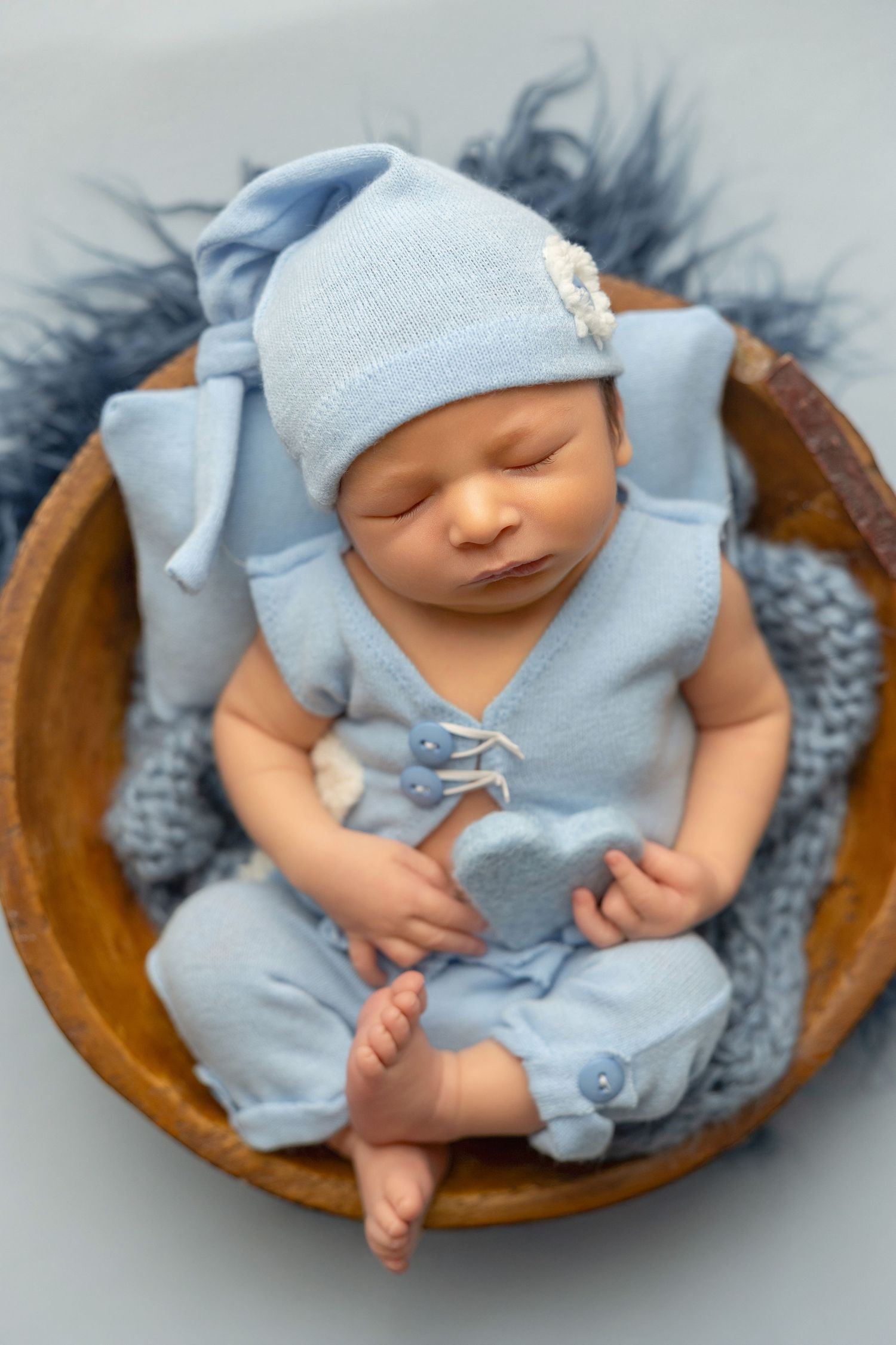 Sleeping newborn baby in light blue outfit rests peacefully in wooden bowl with gray blanket.