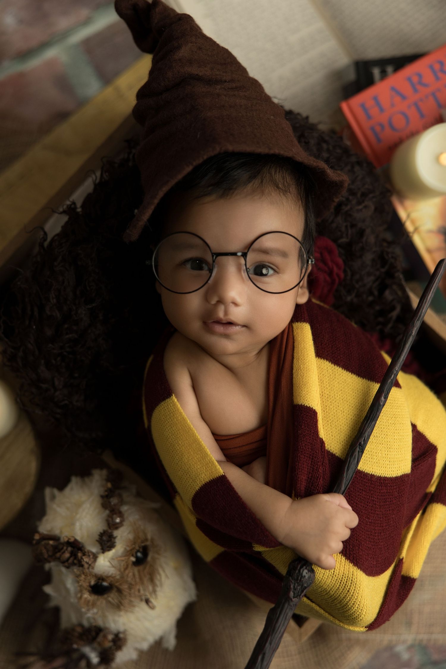 Young Harry Potter fan dressed in Gryffindor scarf and brown wizard hat holds a wand while wearing round glasses.
