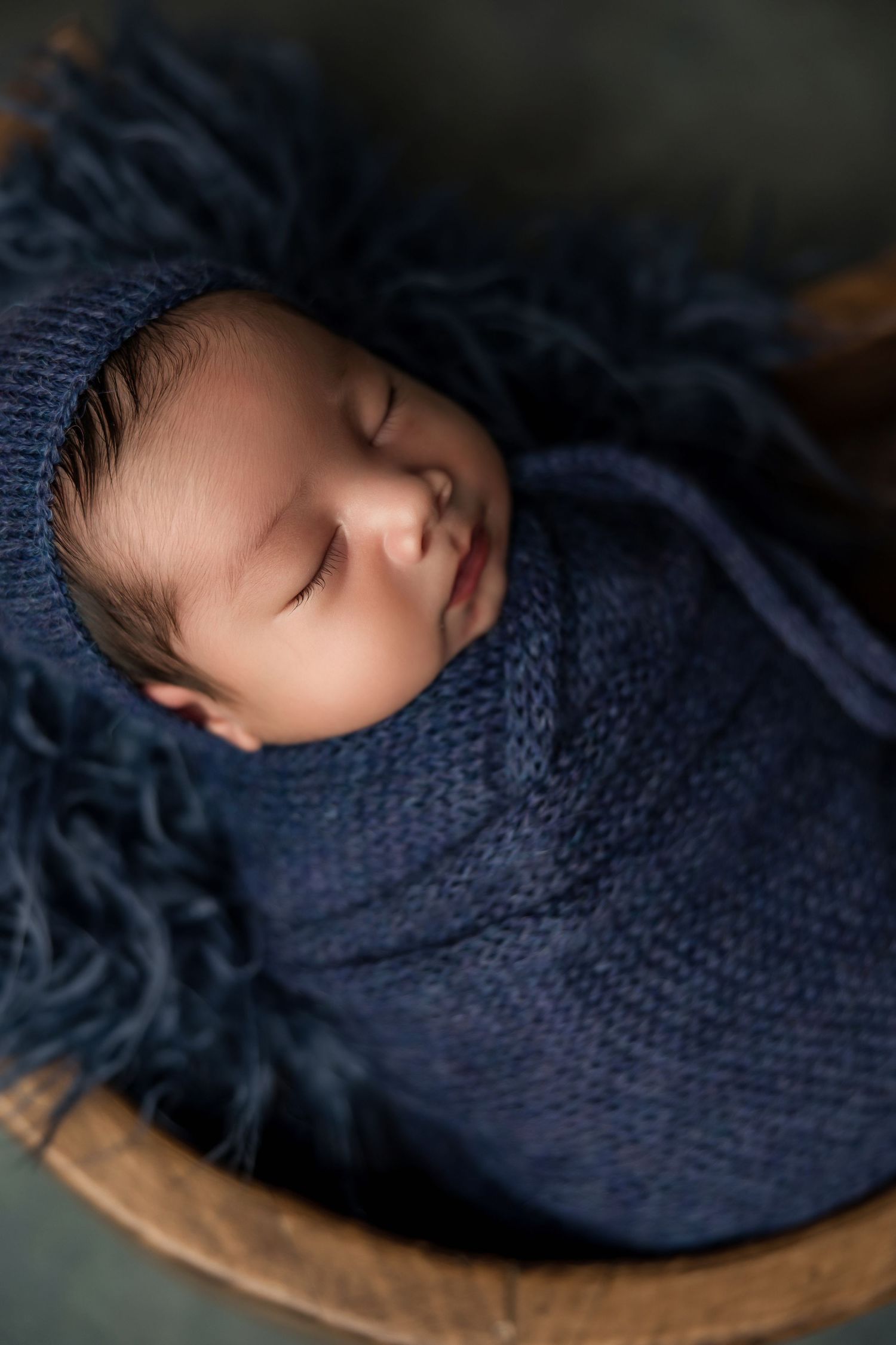 Peaceful sleeping baby wrapped in cozy navy blue knit blanket resting in wooden bowl.