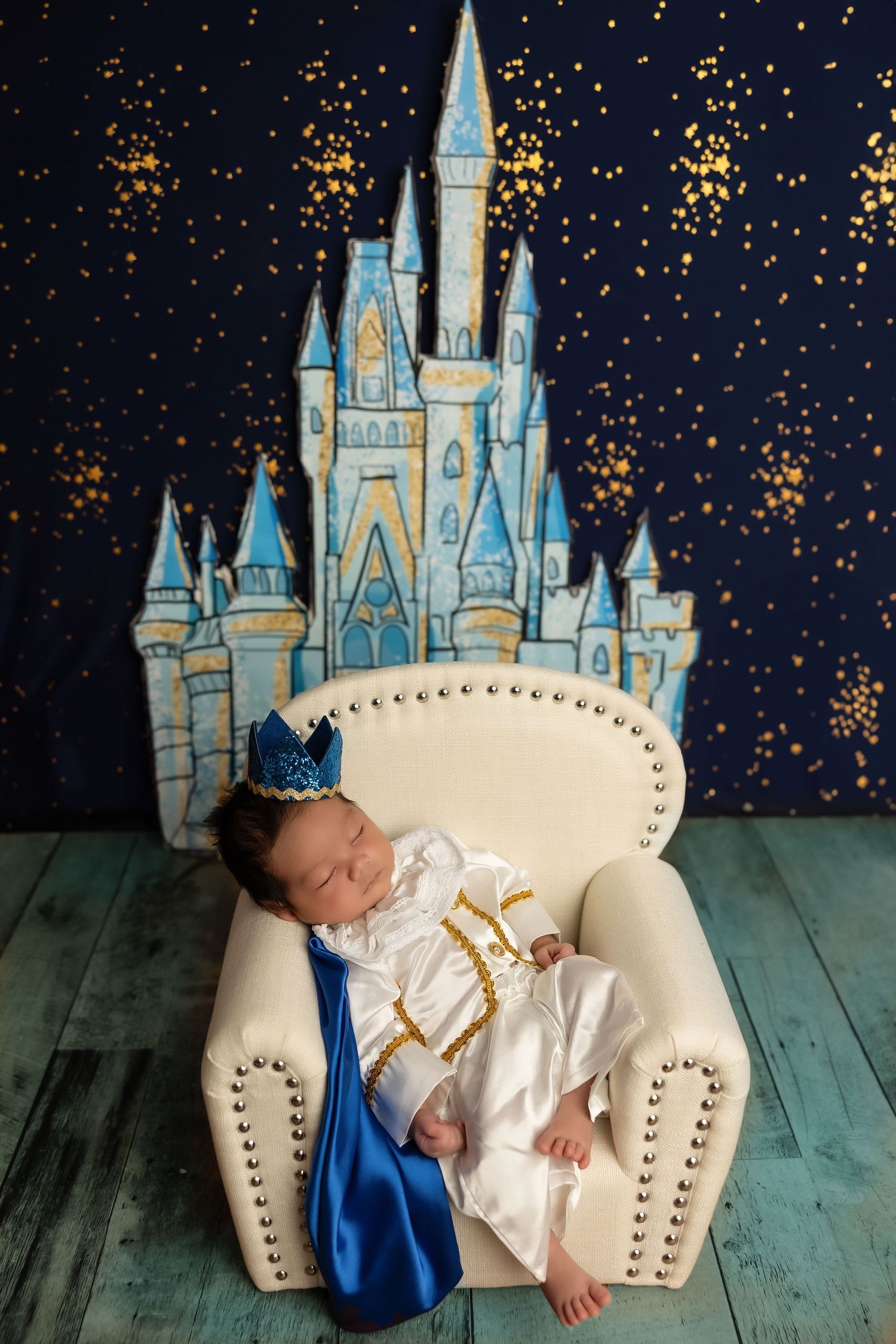 A sleeping baby in white dress lies peacefully on a cream chair against a backdrop of a fairy tale castle and starry night sky.