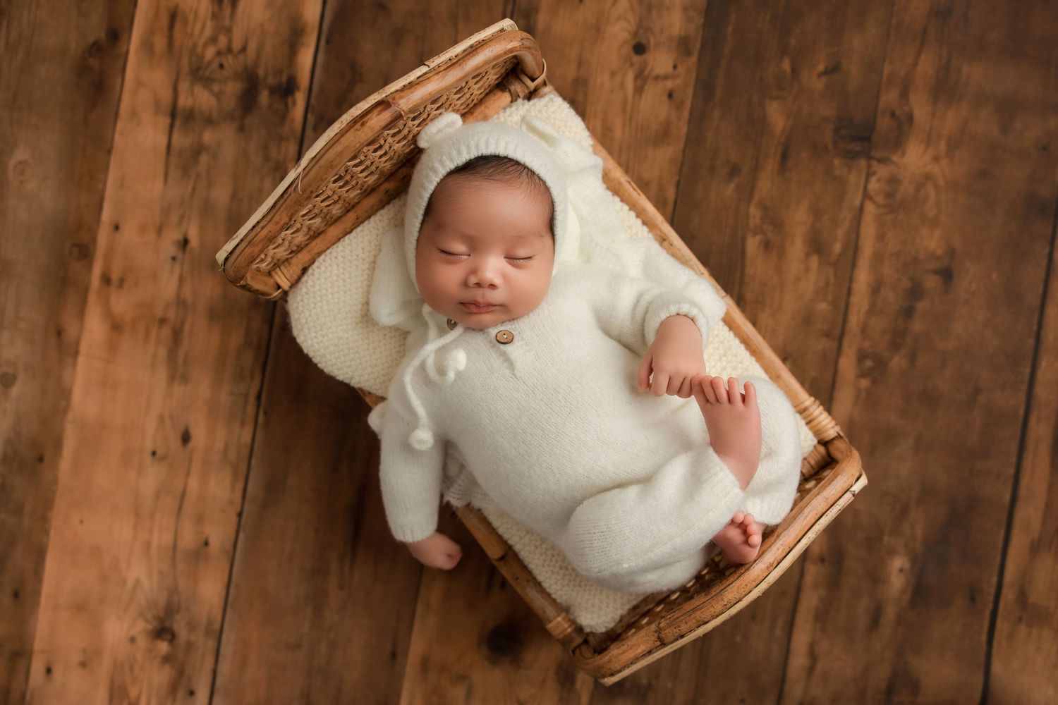 Newborn baby in white outfit sleeping peacefully in wooden basket against rustic wood background.