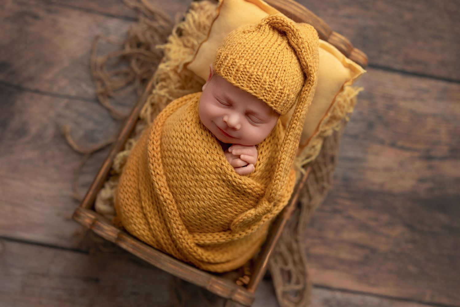 A small baby wearing a cozy yellow knit bonnet lies sleeping on a rustic wooden surface.