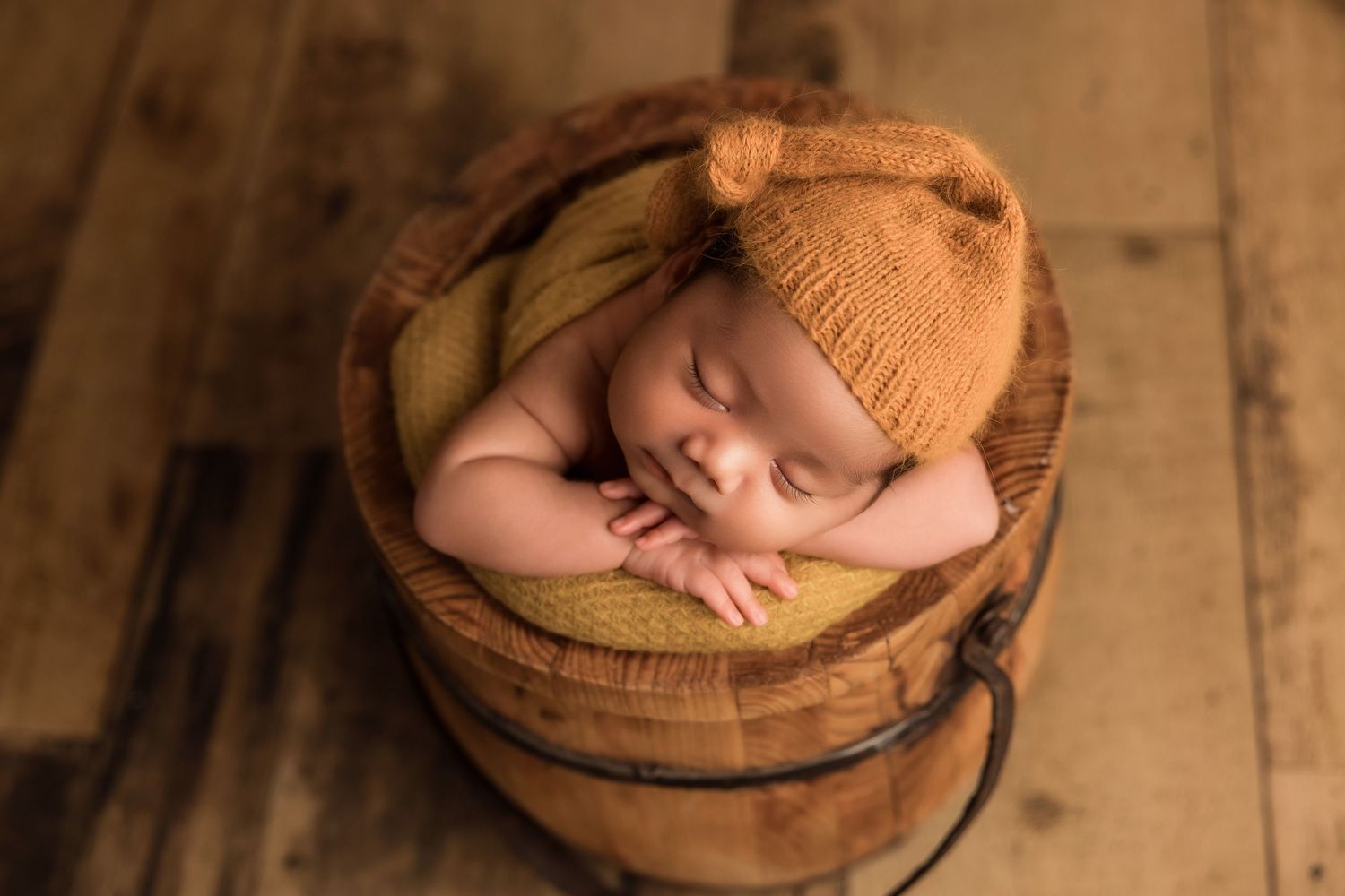 A sleeping newborn baby wearing a yellow bonnet lies peacefully in a rustic wooden bowl on a wooden surface.