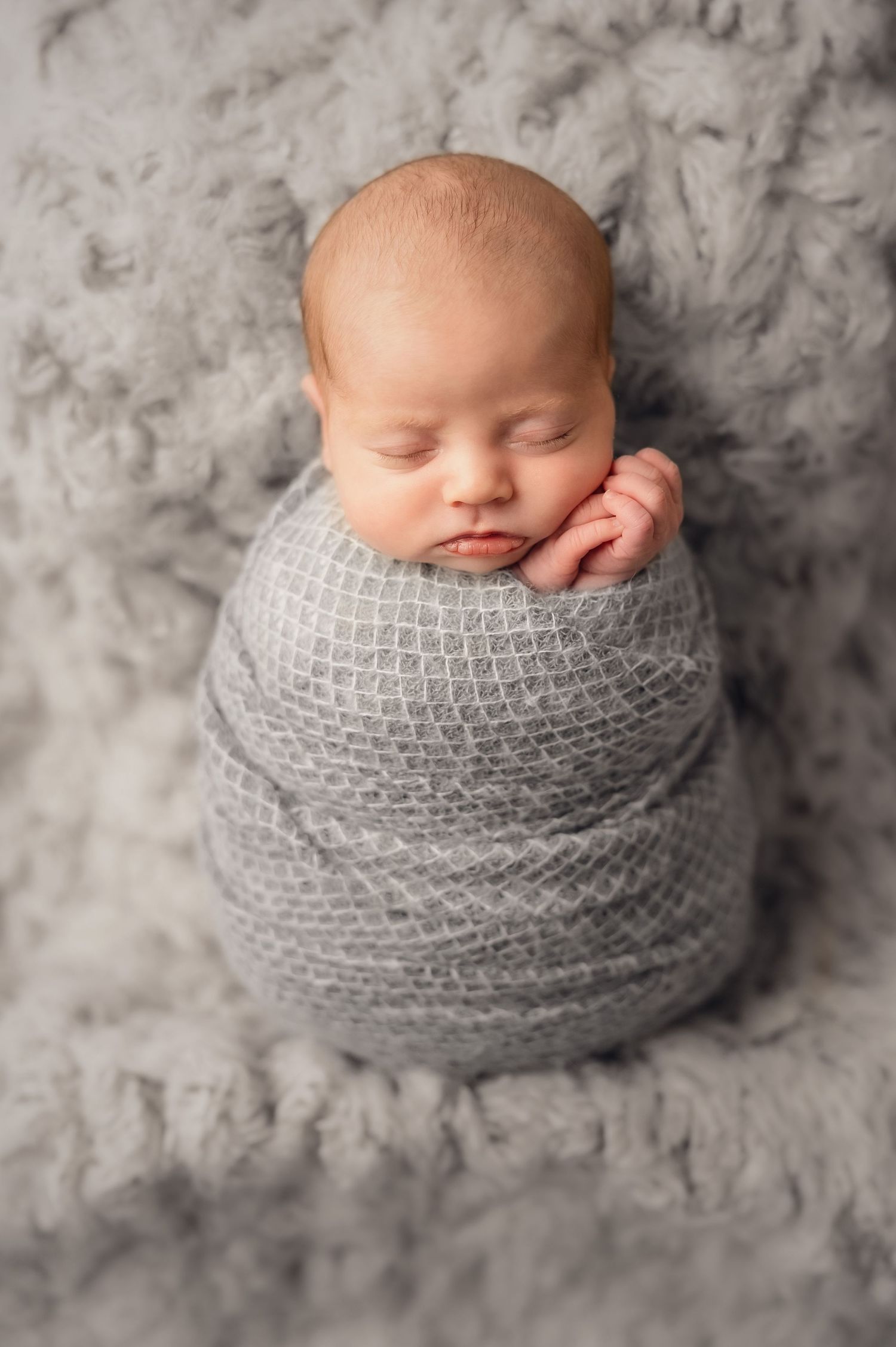 Sleeping newborn baby swaddled in soft gray knit wrap on textured gray backdrop in professional portrait series.