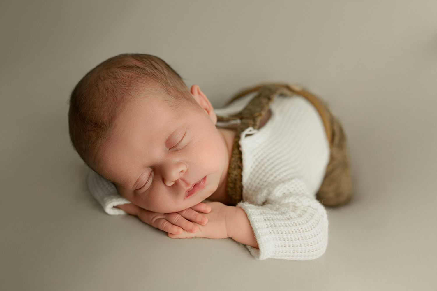 Peaceful sleeping newborn in white knit sweater rests on neutral gray backdrop during professional photo session.