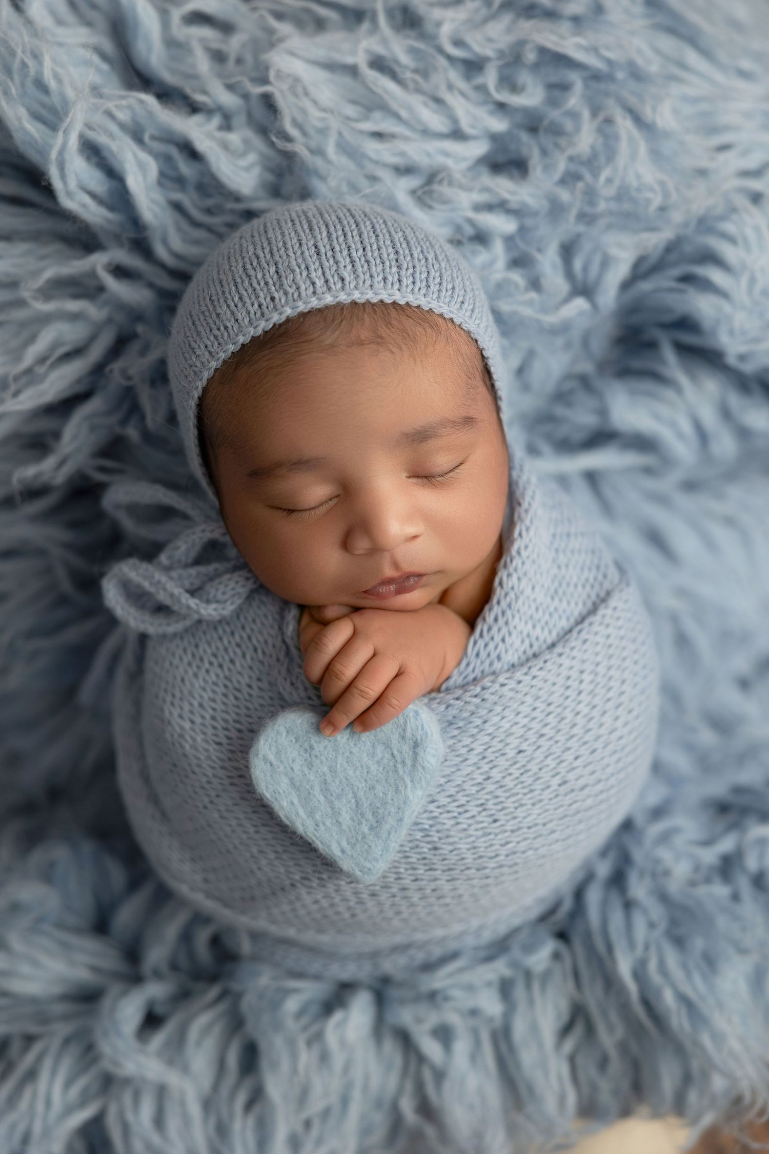 Sleeping newborn baby wrapped in soft blue knit blanket on fluffy gray backdrop in professional photo series.