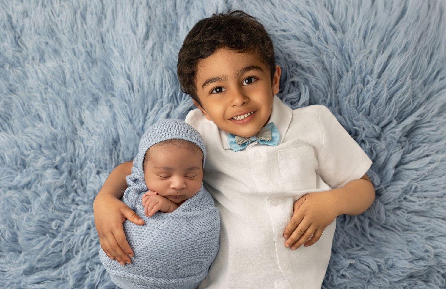 Two siblings pose together on soft blue fur backdrop during newborn photo session.