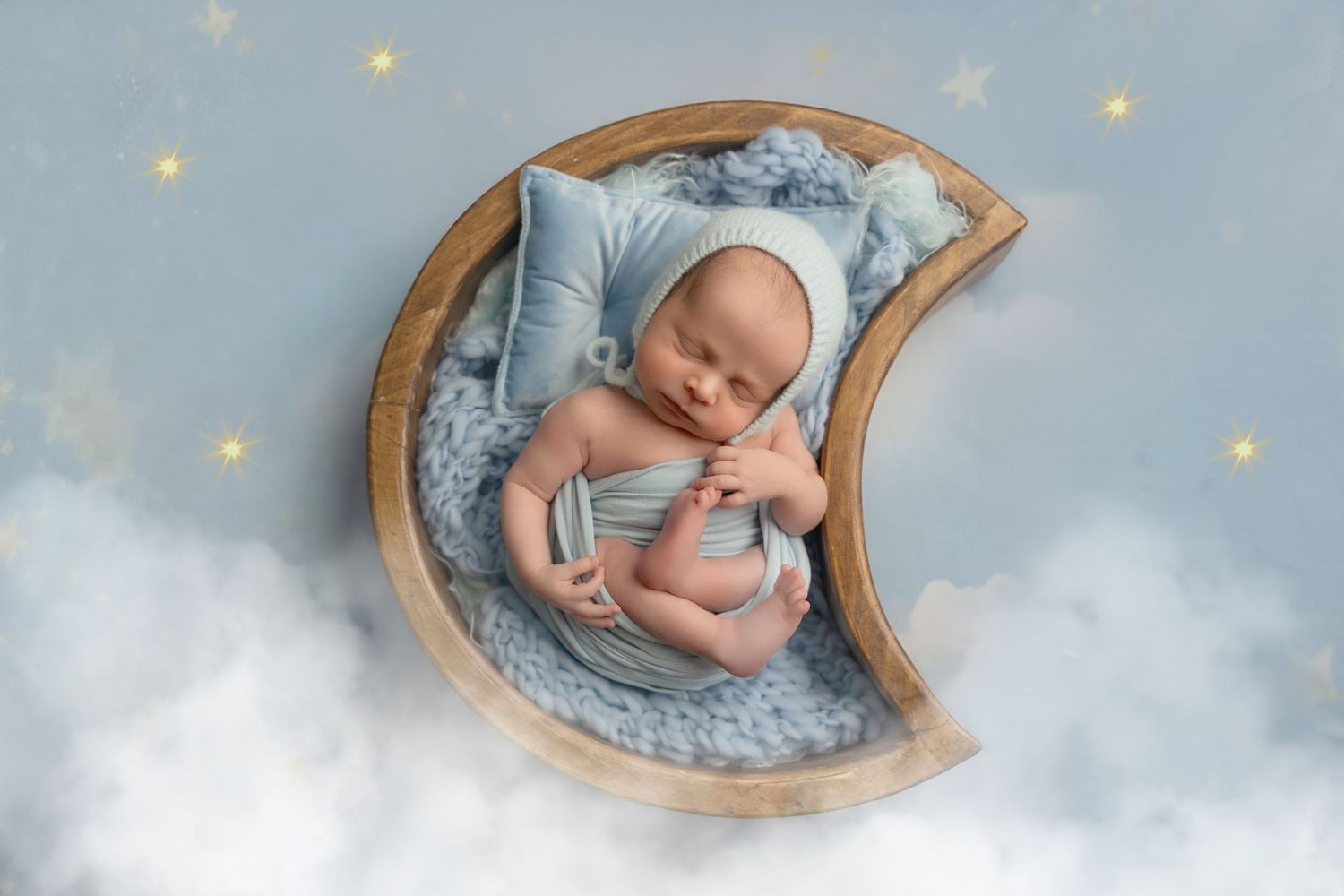 A sleeping newborn rests peacefully on a crescent moon prop with soft blue bedding and twinkling stars in the background.