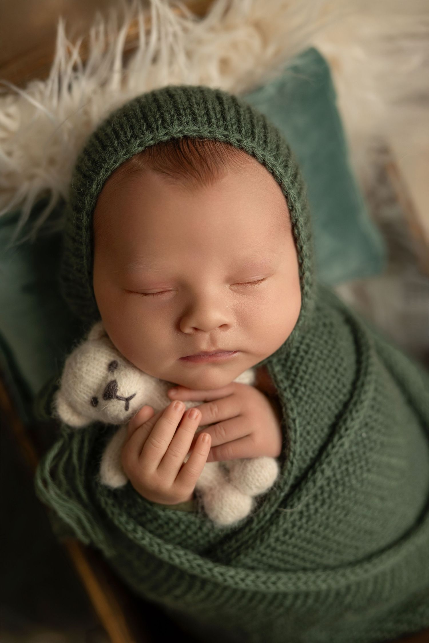 Sleeping newborn wrapped in green knit blanket holding tiny teddy bear during professional photography session.