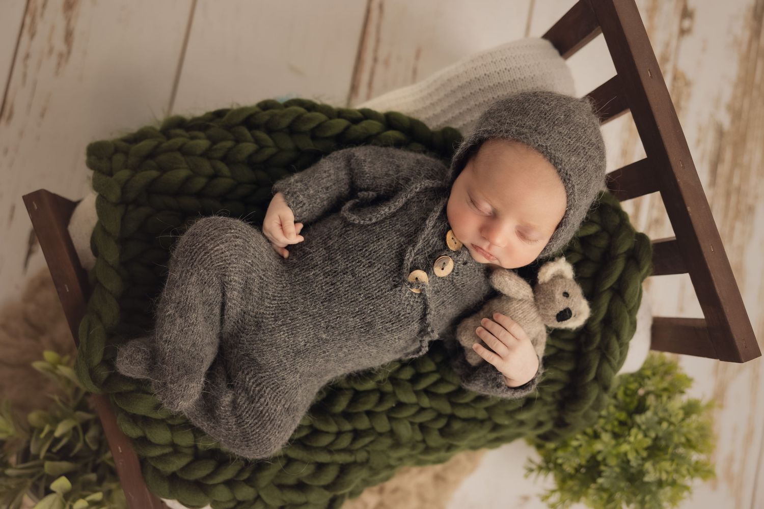 Baby in gray sweater crawling on wooden floor with greenery in background.