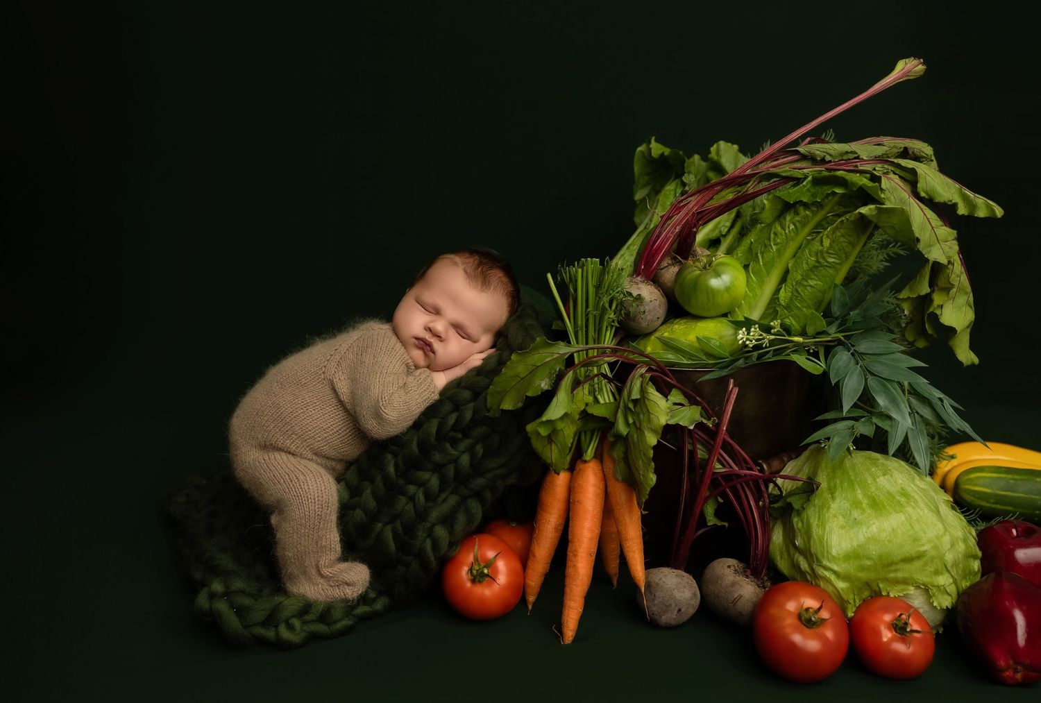 Sleeping baby in brown onesie lies peacefully next to fresh vegetables including carrots, tomatoes, and cabbage on dark backdrop.