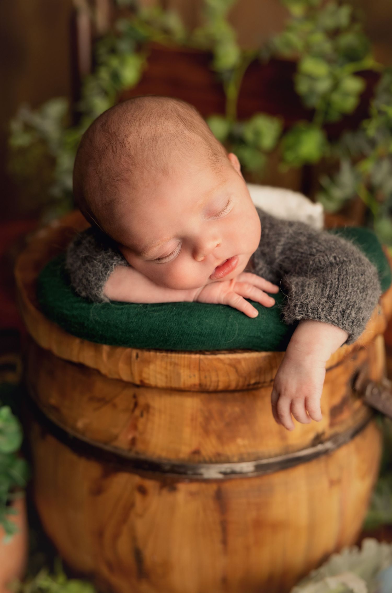 Sleeping newborn in gray wrap nestled in rustic wooden barrel with green foliage backdrop.