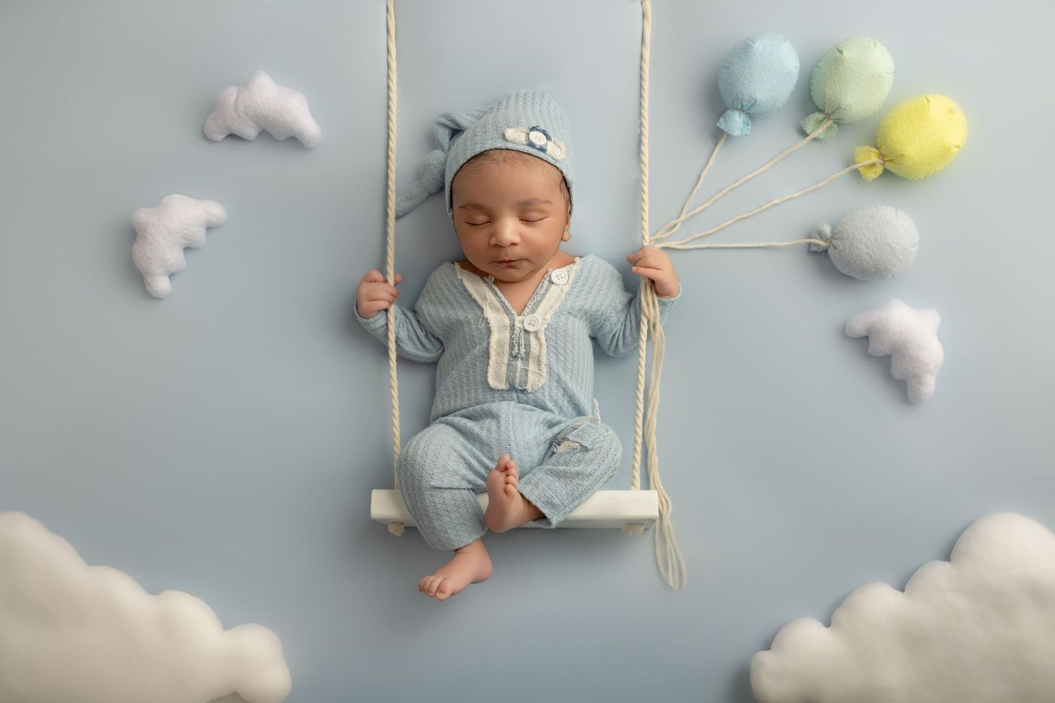 A sleeping baby on a wooden swing surrounded by fluffy white clouds and pastel pom poms against a blue background.