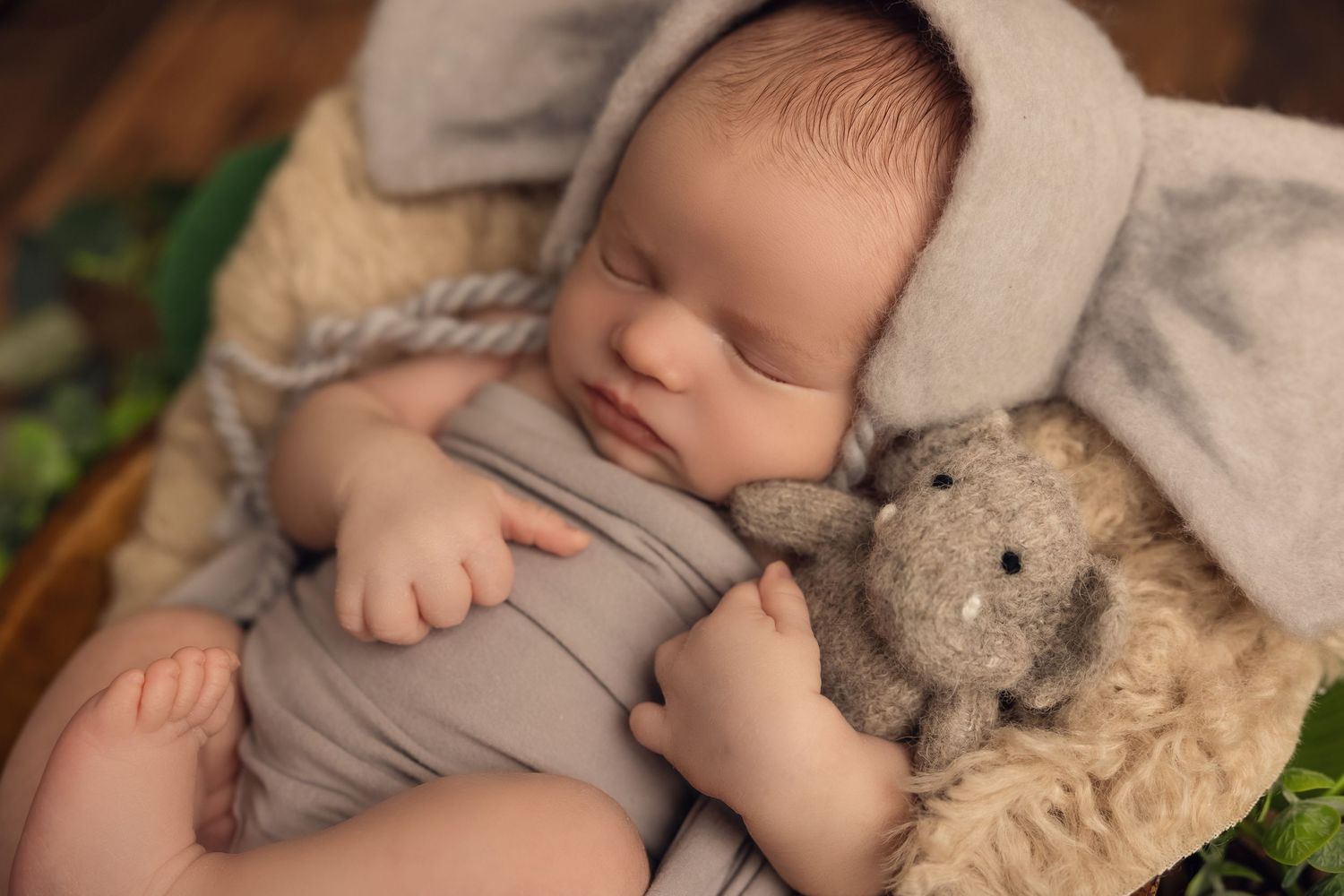 Sleeping newborn in gray wrap and bonnet rests peacefully next to plush teddy bear on soft fur blanket.