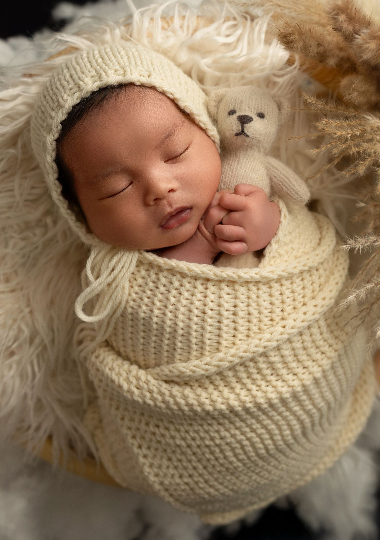Peaceful sleeping infant wrapped in cream knitted blanket with small teddy bear peeking out against fluffy white backdrop.