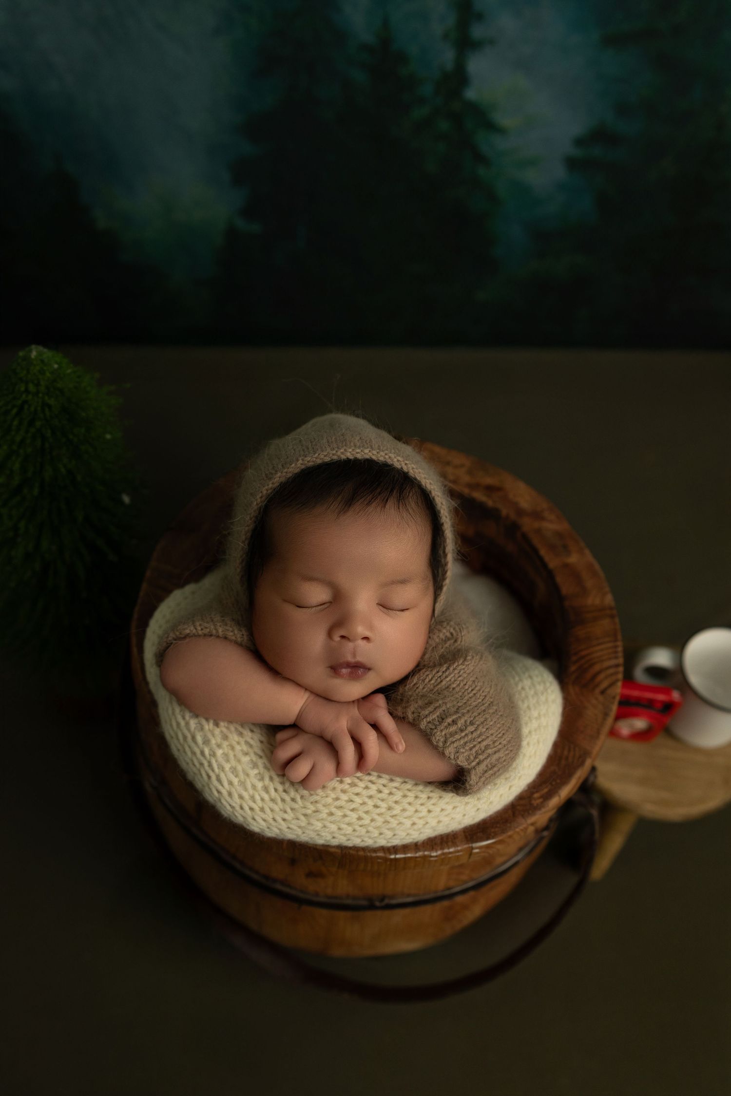 A sleeping newborn in a cozy knit outfit rests peacefully in a wooden bowl against a dark moody backdrop.