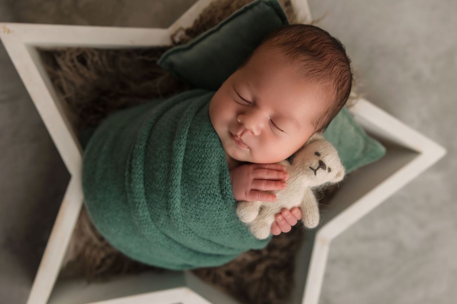 A sleeping newborn baby wearing a green sweater lies peacefully on a white star-shaped cushion.