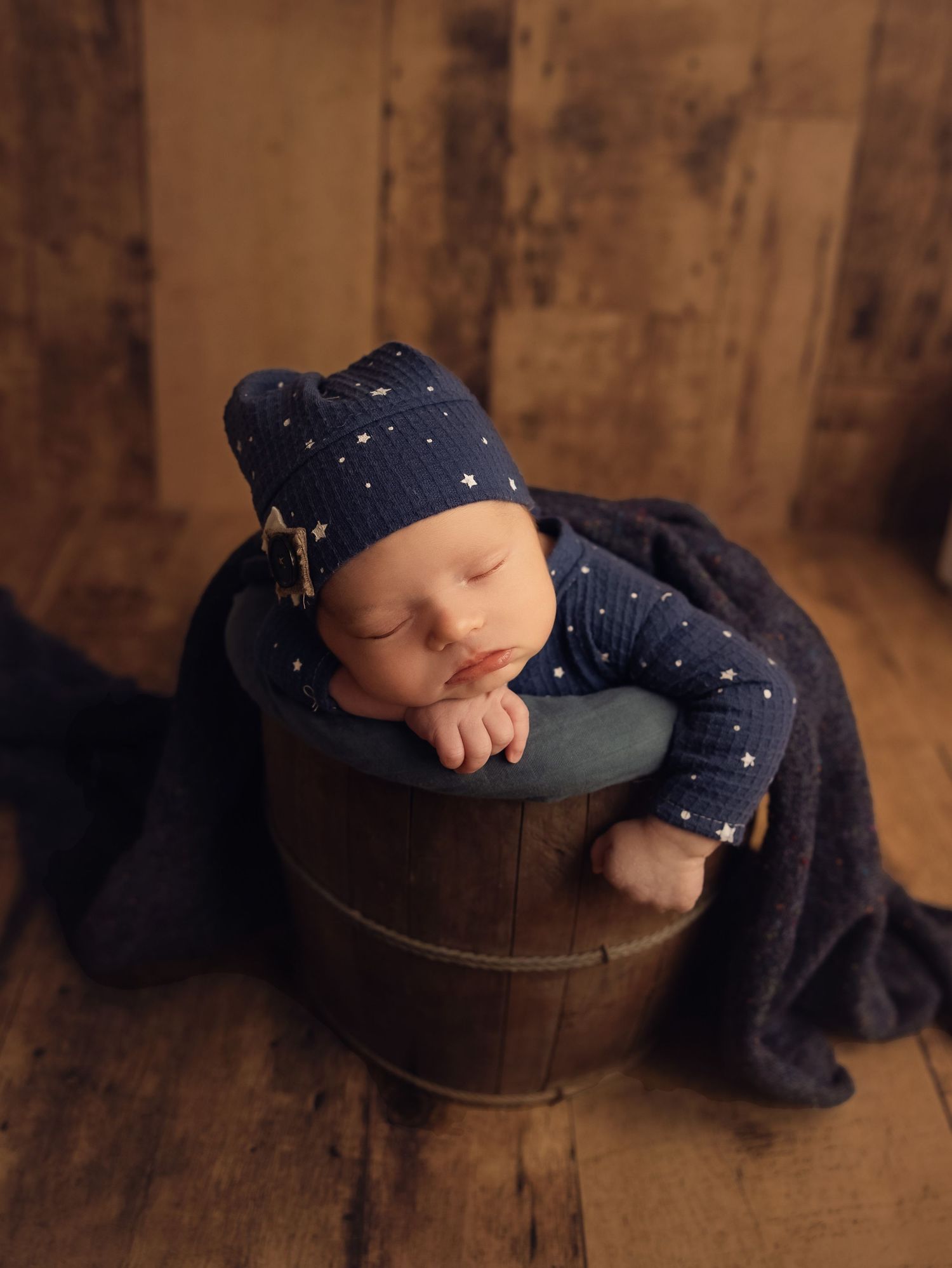 A sleeping baby wearing a navy polka dot outfit rests peacefully in a rustic wooden bucket on a hardwood floor.
