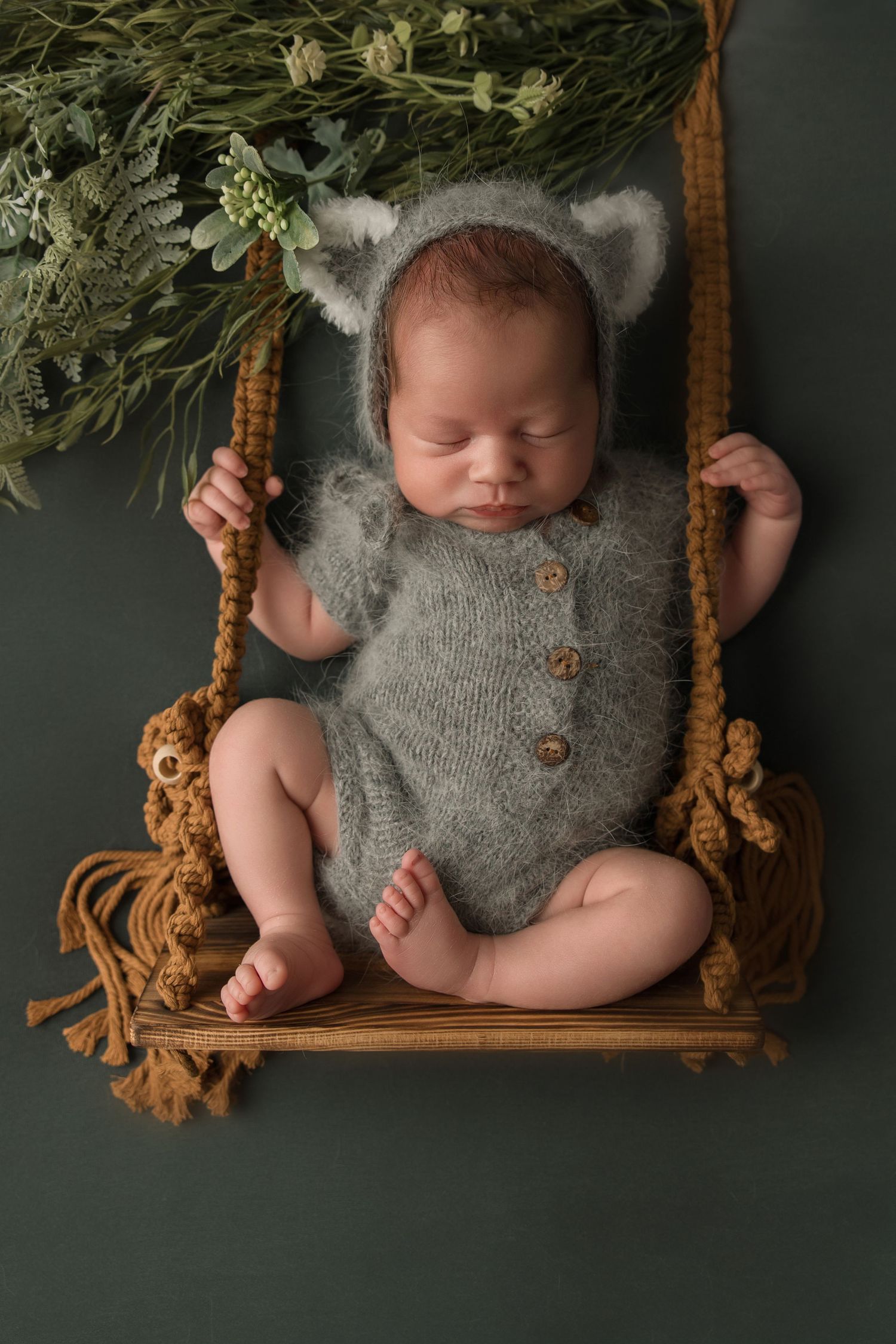 Sleeping newborn wearing gray knit romper with animal ears rests peacefully on wooden swing with greenery backdrop.