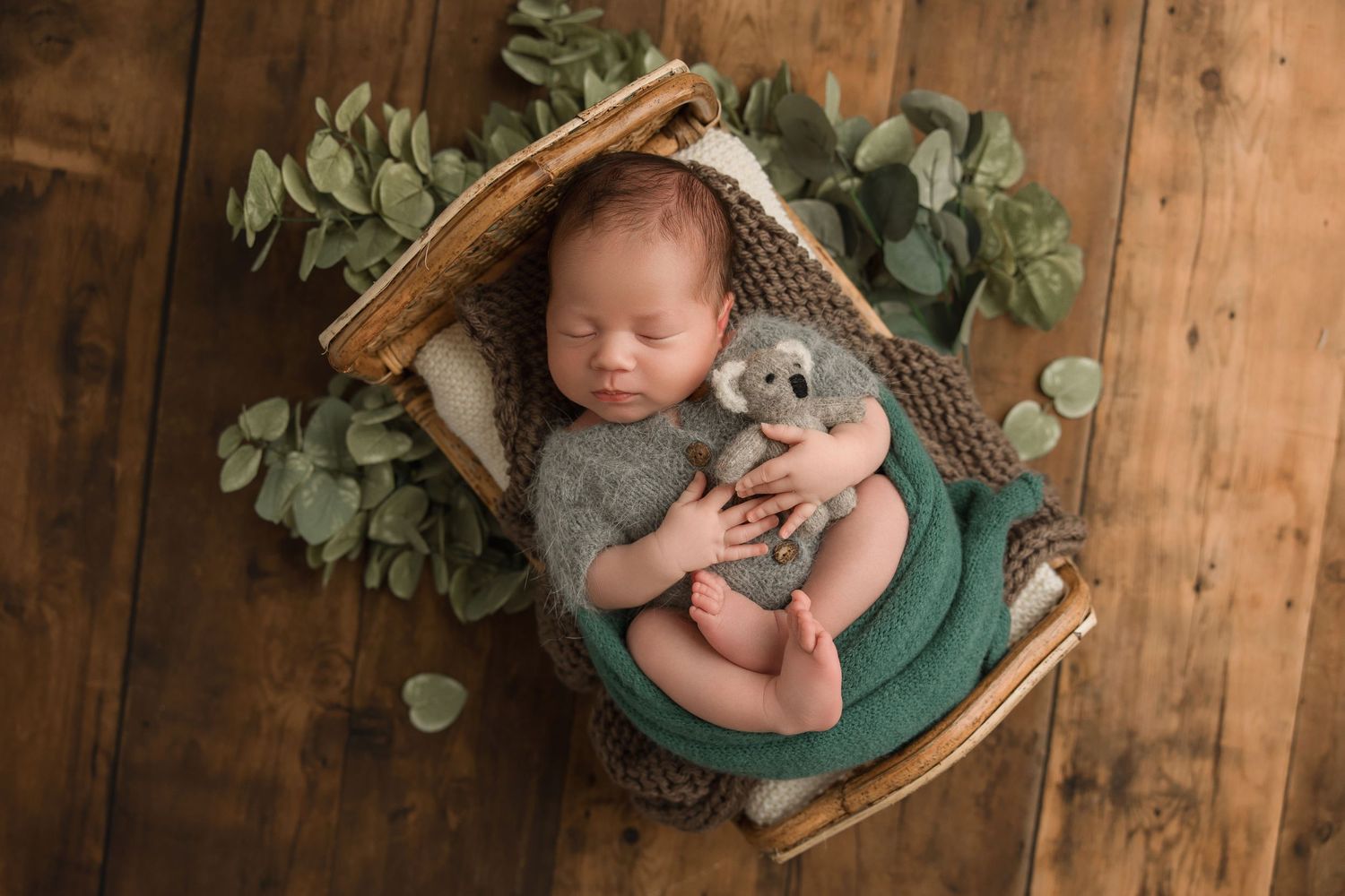 Newborn baby sleeping peacefully in woven basket with eucalyptus leaves and teal blanket on wooden floor.