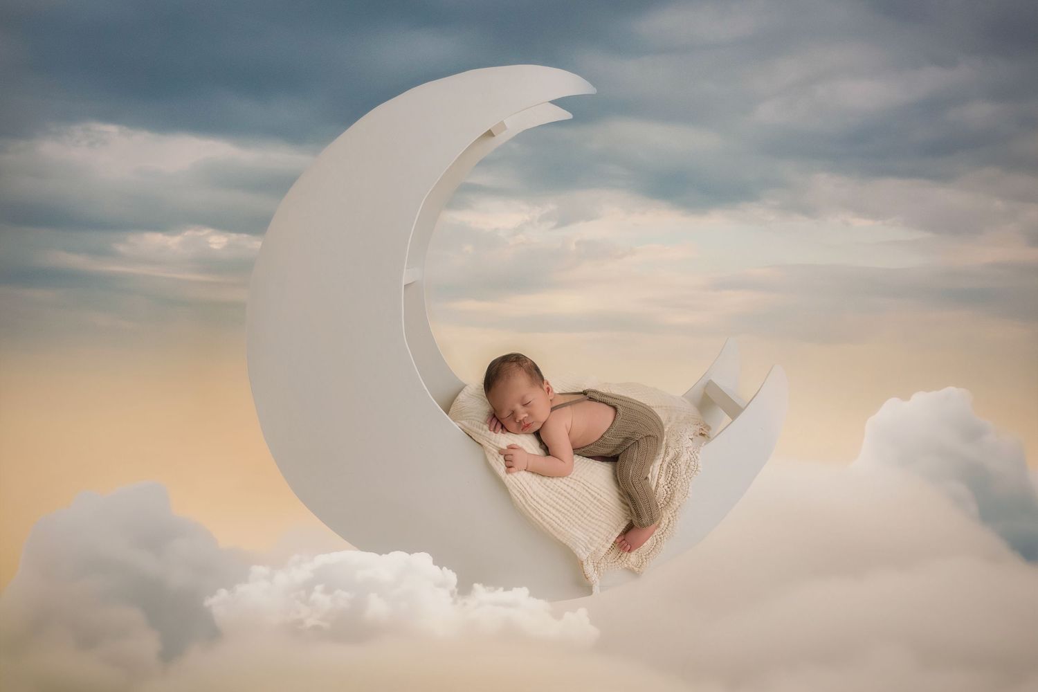 A sleeping baby rests peacefully on a decorative crescent moon prop among dreamy clouds at sunset.