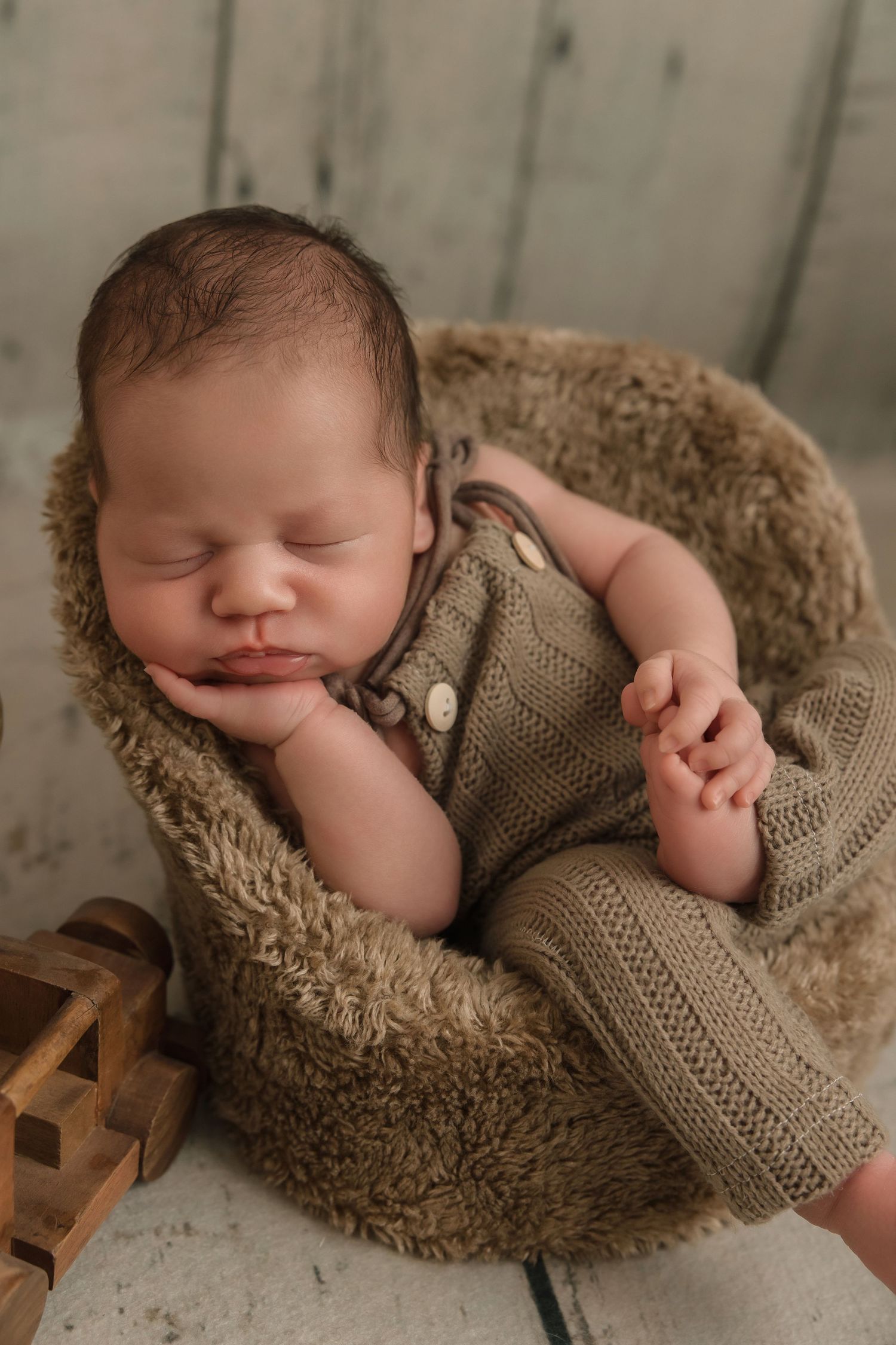 Sleeping newborn wearing knit romper rests peacefully in cozy brown furry nest during professional photo session.
