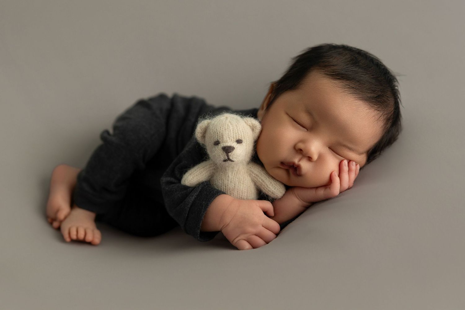Sleeping baby in black outfit cuddling with small white teddy bear on grey background.