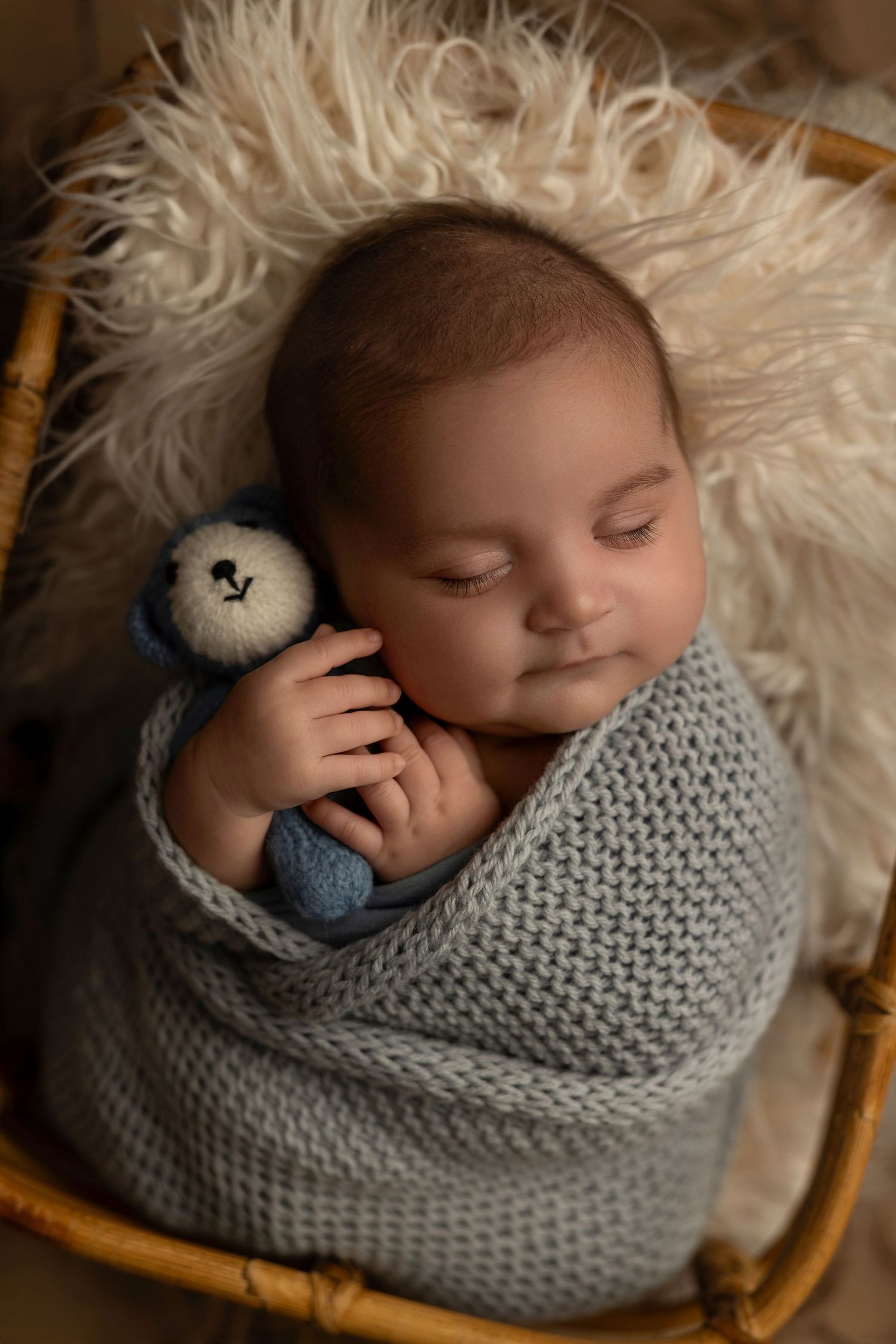 Sleeping baby wrapped in gray knit blanket with small stuffed toy on fluffy white fur background.