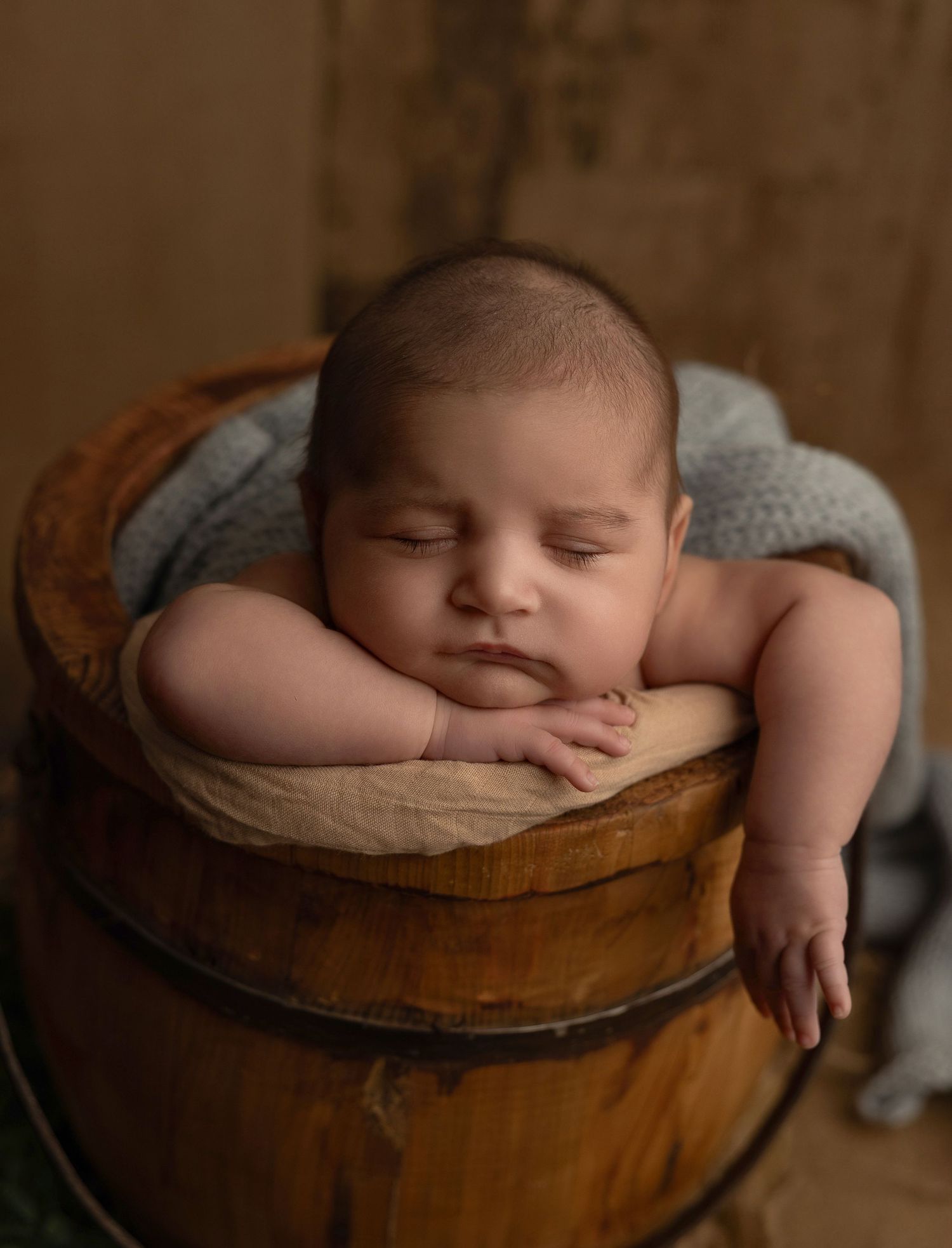 A sleeping newborn rests peacefully in a wooden bowl lined with soft beige fabric during a professional photography session.