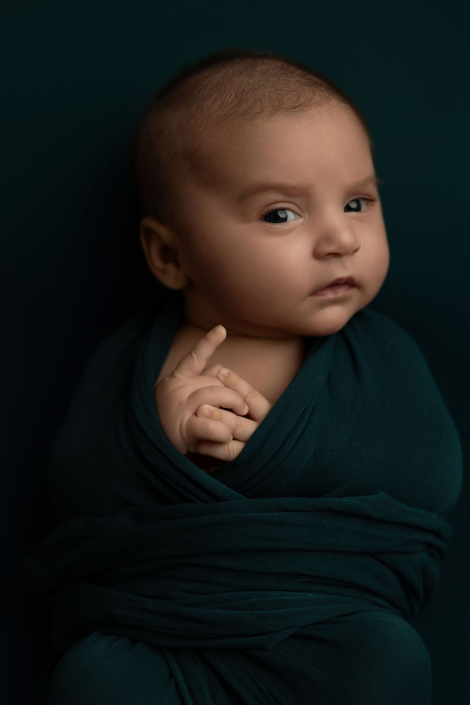 A series of moody studio portraits showing a baby wrapped in dark teal fabric against a black background.