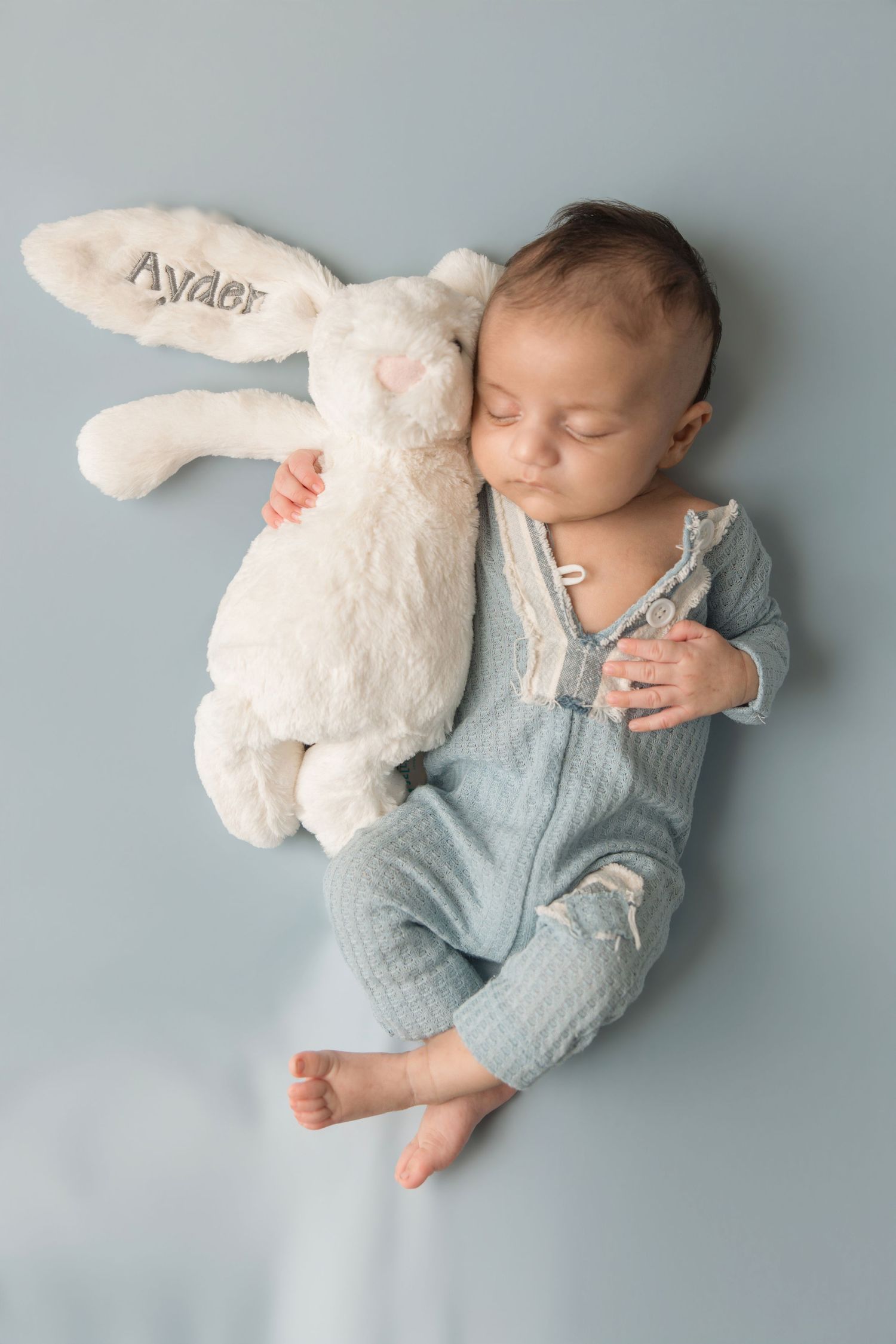 Sleeping newborn in light blue sleeper rests peacefully with white plush bunny on gray backdrop.