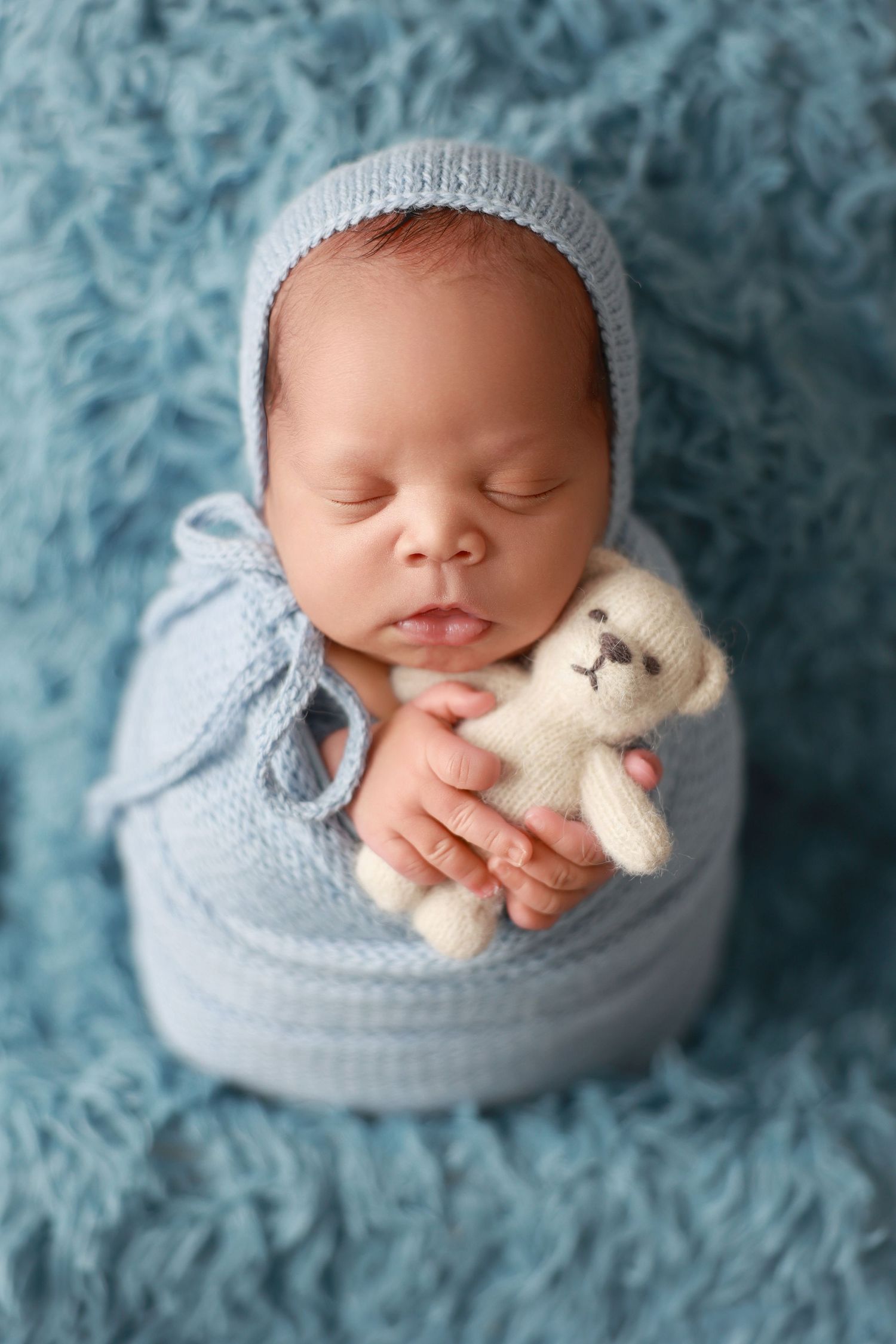 Sleeping newborn wrapped in soft blue blanket with small white teddy bear on textured blue background.