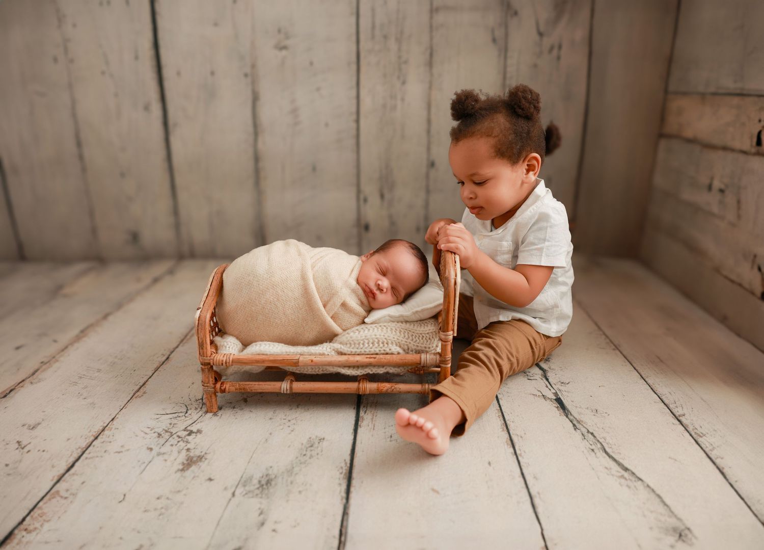 A toddler in brown pants sits next to a tiny wooden bed with a sleeping baby on a rustic wooden floor.