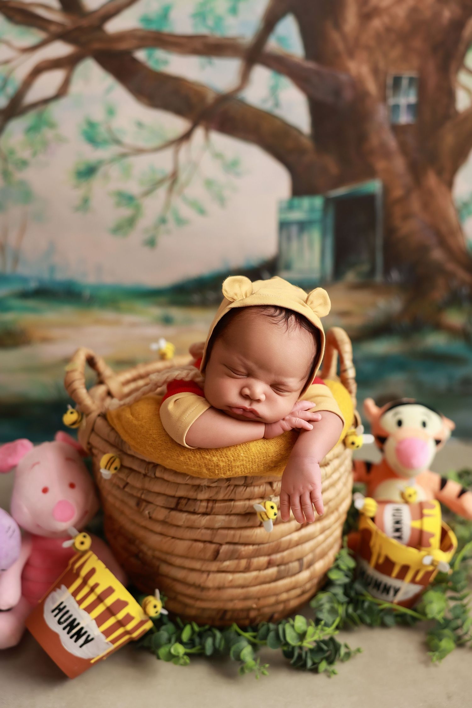 Sleeping baby in yellow outfit nestled in honey pot basket with Winnie the Pooh themed stuffed animals and decorations.