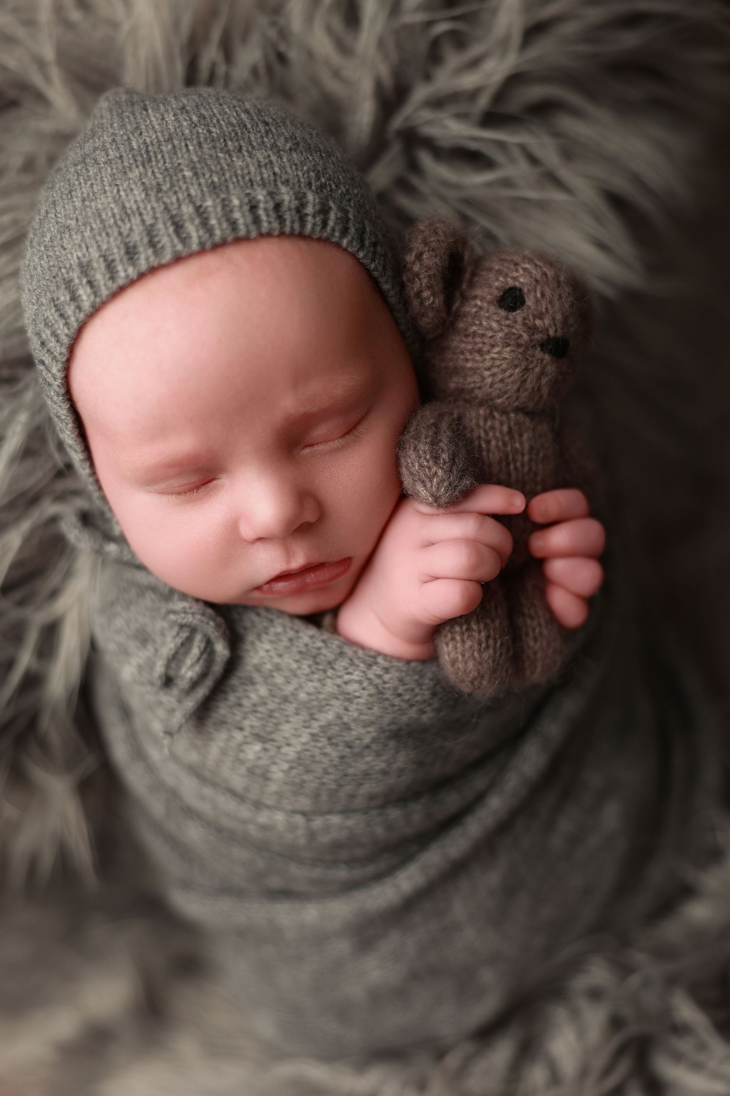 Sleeping newborn wrapped in gray knit swaddle with matching hat and small teddy bear on furry backdrop.