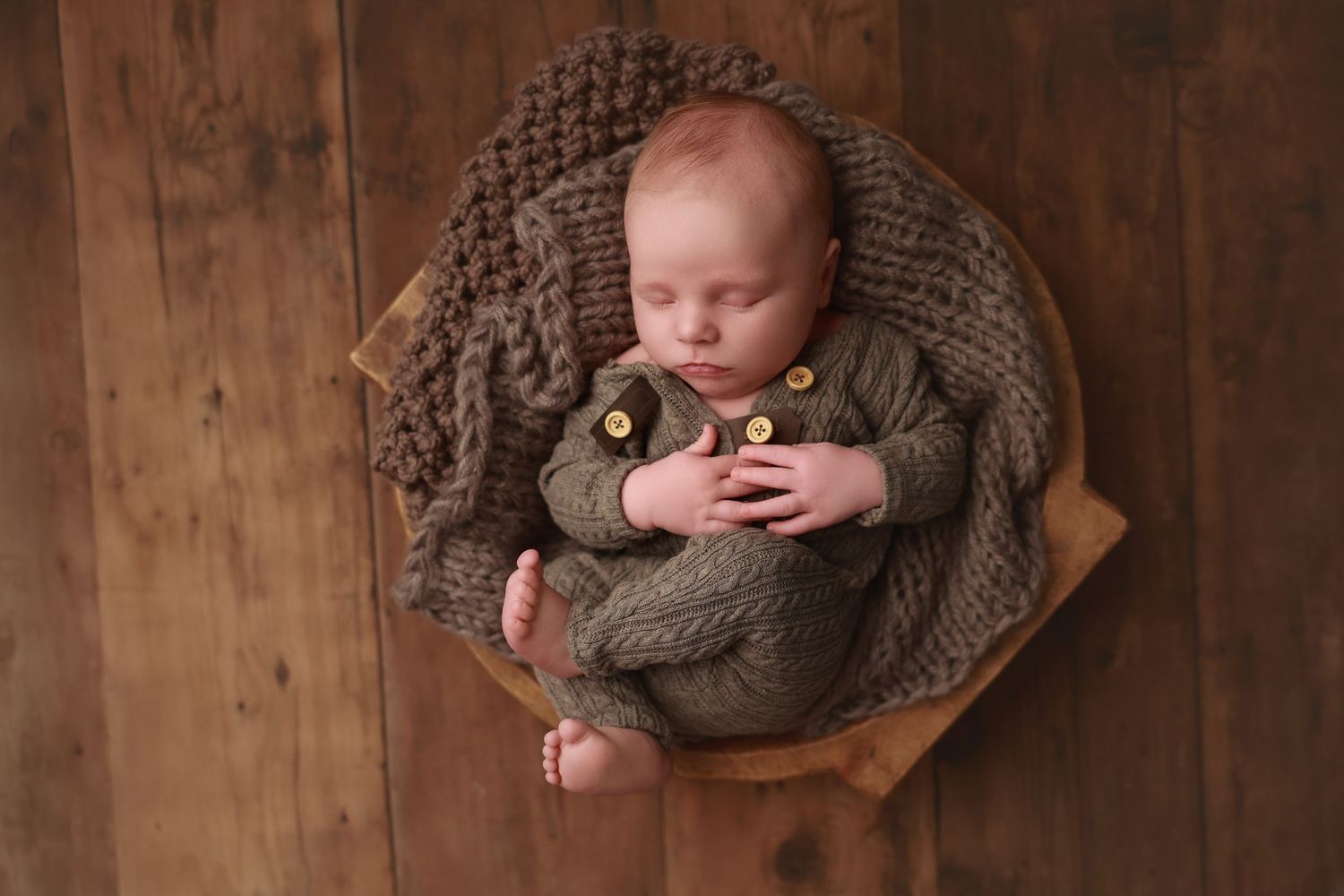 Sleeping baby wrapped in cozy brown knit blanket lying in wooden bowl against rustic wooden background.