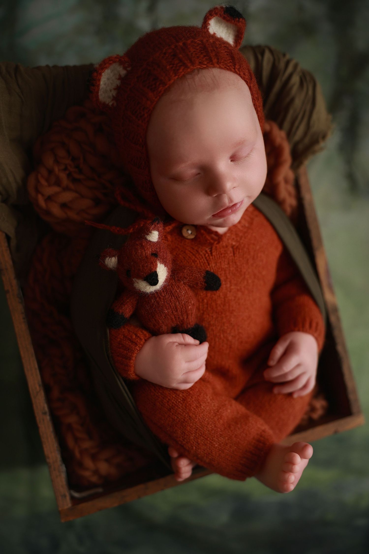 A small baby wearing a cozy fox-themed orange onesie sleeps peacefully in a wooden basket.