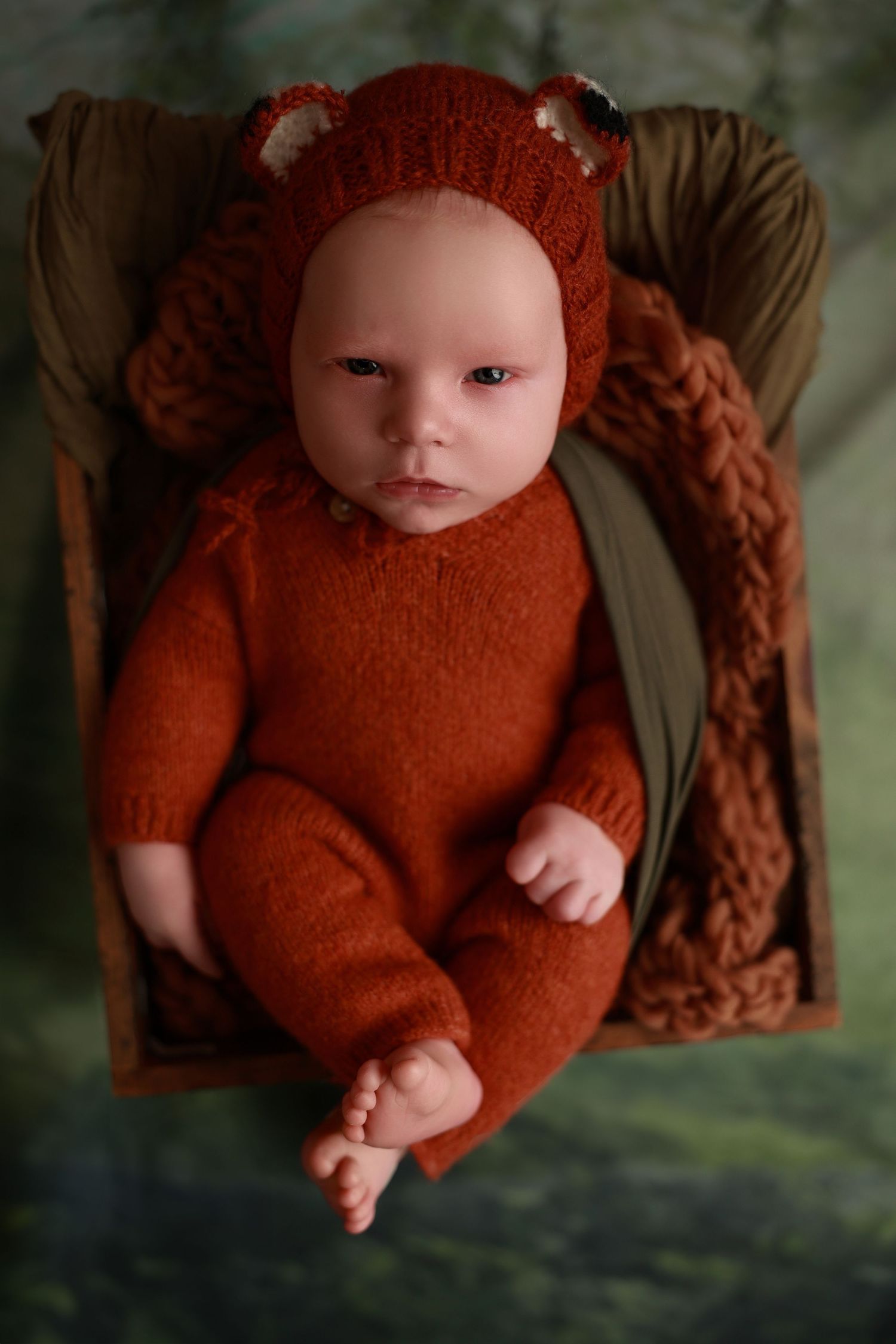 Infant wearing rusty orange knit bear outfit with ears lying on brown cushioned surface.