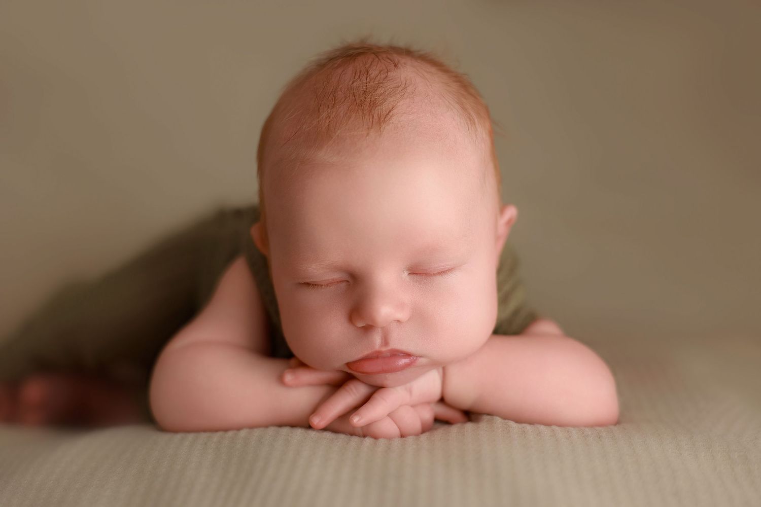 A sleeping newborn baby rests peacefully with folded arms on a beige backdrop in soft lighting.
