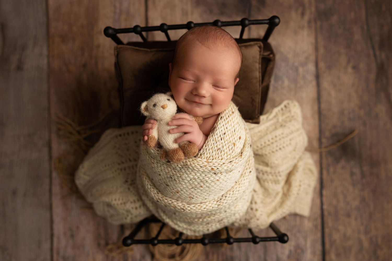 A newborn baby wrapped in a cream-colored knit blanket lies peacefully on a dark wooden backdrop.
