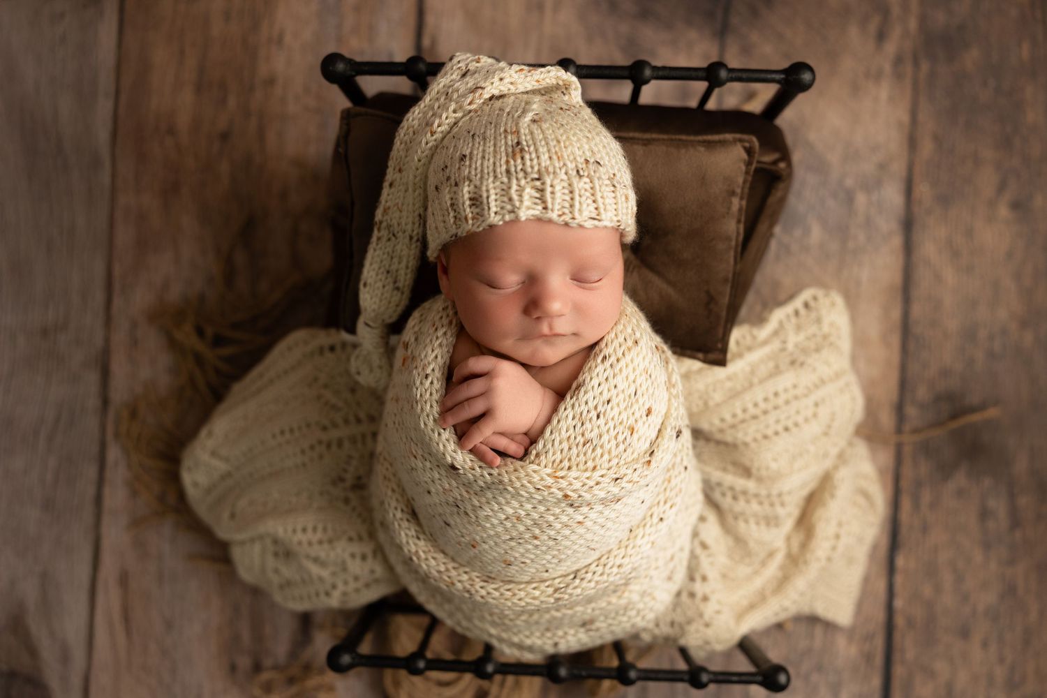 Sleeping newborn wrapped in cream knit blanket wearing matching hat rests peacefully in rustic wooden basket.
