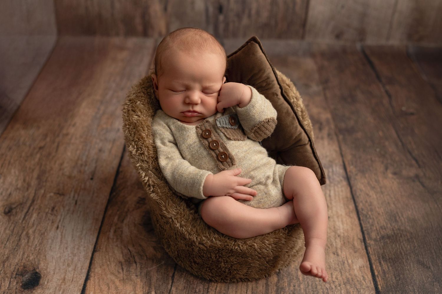 Sleeping baby wearing cream knit romper rests peacefully in brown woven basket on rustic wooden floor.